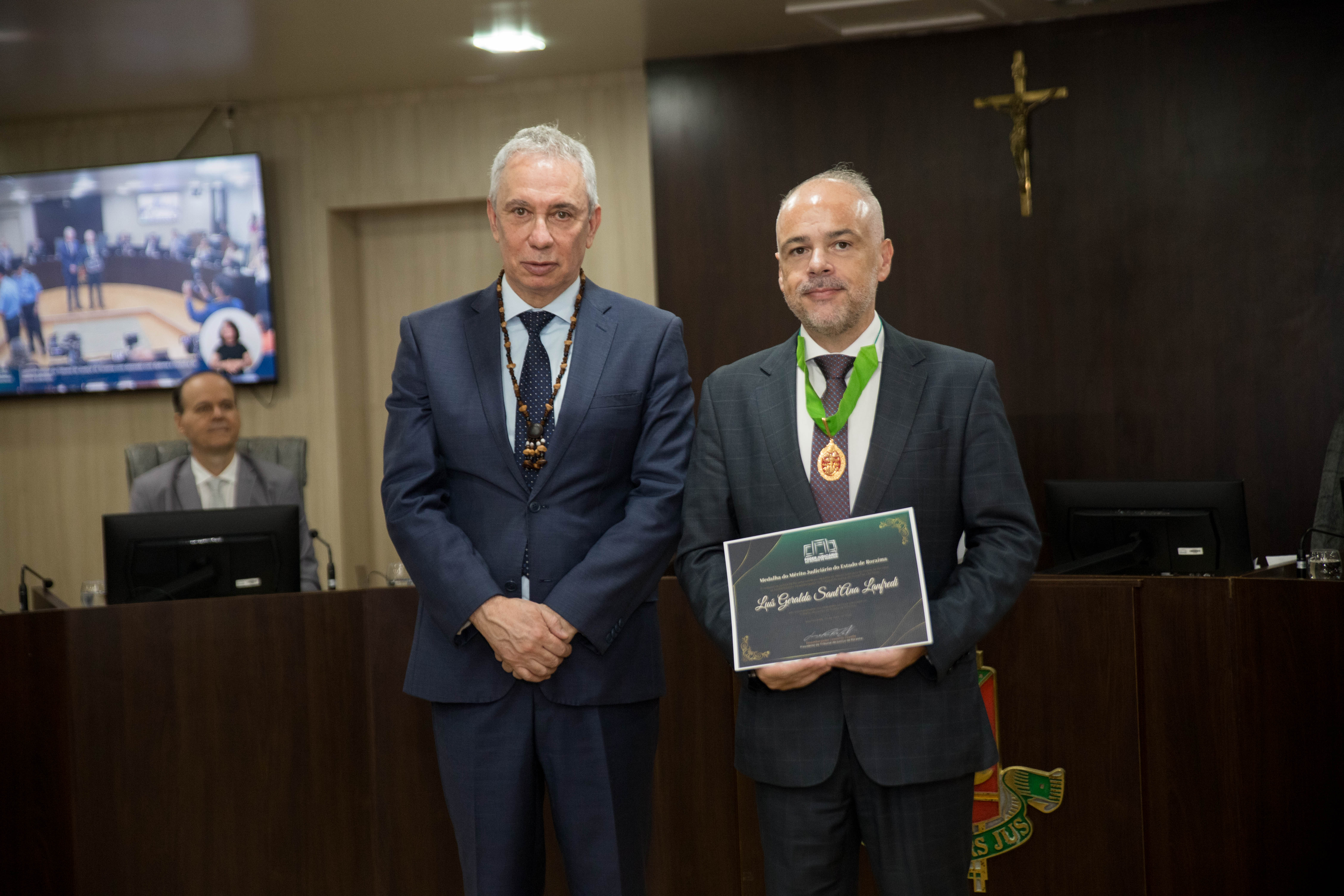 A imagem colorida mostra o Desembargador Luís Lanfredi segurando a Medalha do Mérito Judiciário do Estado de Roraima ao lado do vice-presidente do Tribunal de Justiça de Roraima e coordenador do GMF, Desembargador Almiro Padilha, A imagem colorida mostra o Desembargador Luís Lanfredi segurando a Medalha do Mérito Judiciário do Estado de Roraima ao lado do vice-presidente do Tribunal de Justiça de Roraima e coordenador do GMF, Desembargador Almiro Padilha,