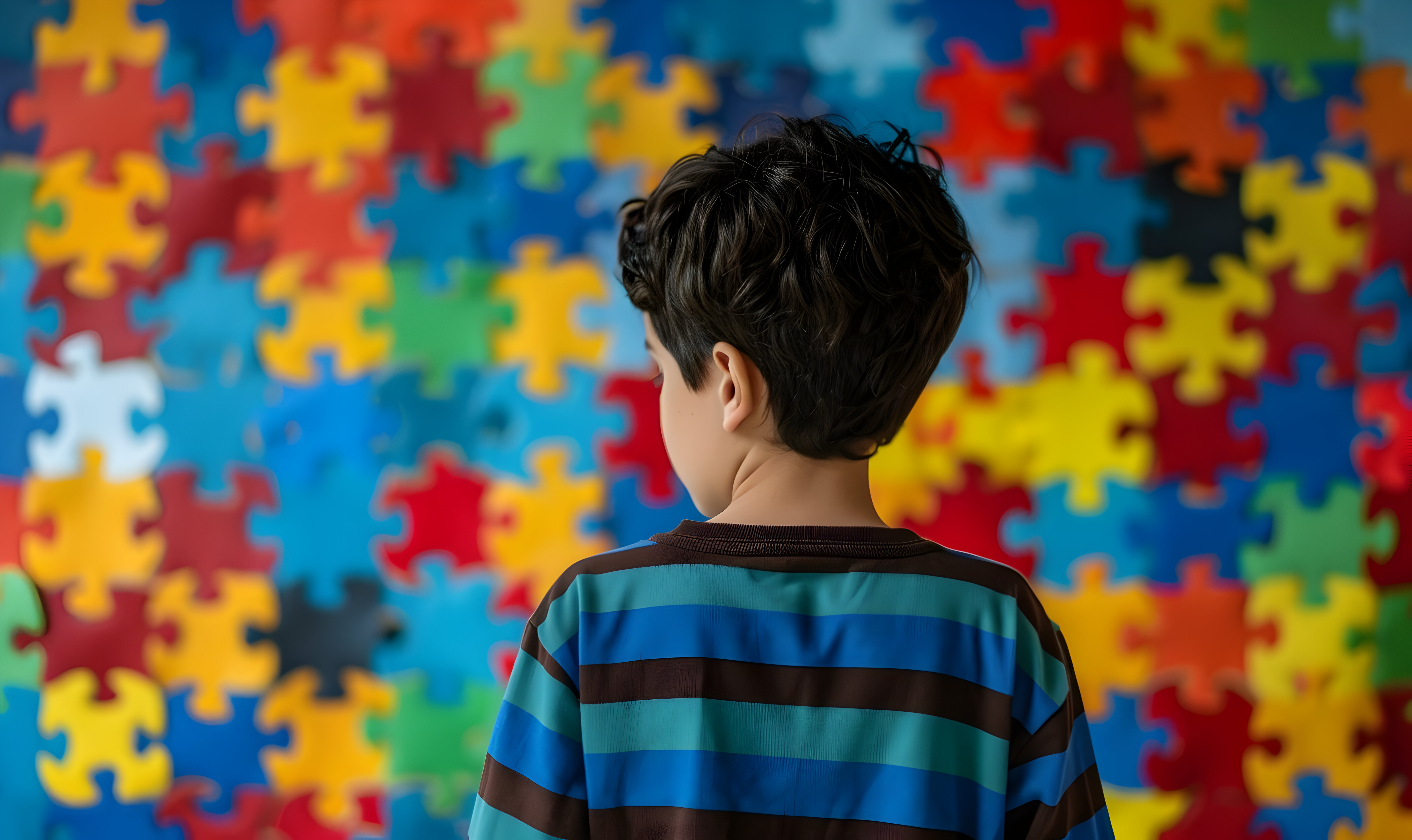 A imagem apresenta um menino está de costas, olhando para uma parede coberta com peças de quebra-cabeça grandes e coloridas. O menino tem cabelo castanho escuro, curto e ondulado, e está vestindo uma camiseta com listras horizontais azuis, verdes e marrons. As peças do quebra-cabeça ao fundo são de várias cores vibrantes, como vermelho, amarelo, azul e verde, preenchendo todo o espaço da imagem A imagem apresenta um menino está de costas, olhando para uma parede coberta com peças de quebra-cabeça grandes e coloridas. O menino tem cabelo castanho escuro, curto e ondulado, e está vestindo uma camiseta com listras horizontais azuis, verdes e marrons. As peças do quebra-cabeça ao fundo são de várias cores vibrantes, como vermelho, amarelo, azul e verde, preenchendo todo o espaço da imagem
