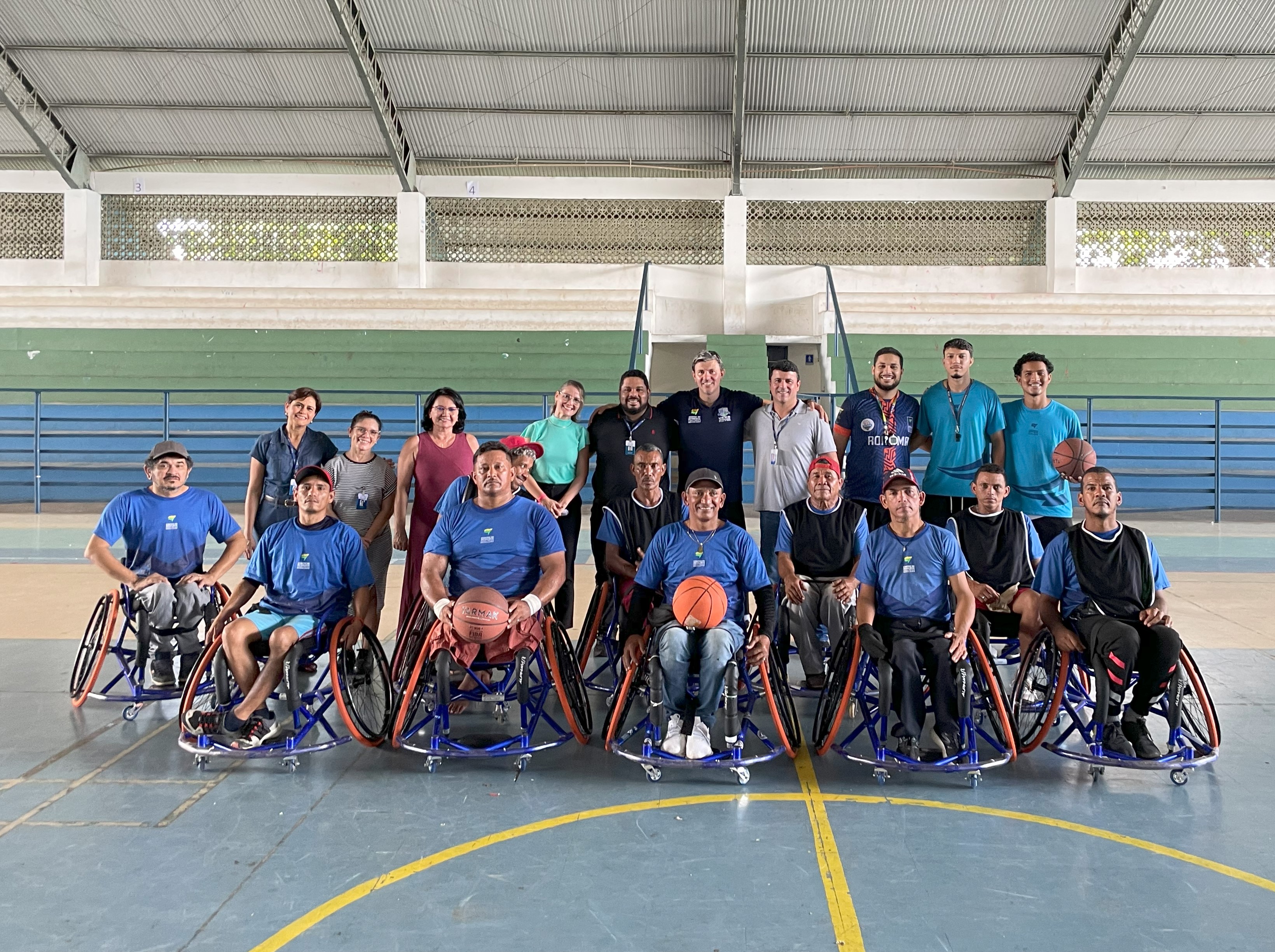 Foto colorida mostra um grupo de aproximadamente 20 pessoas posando para uma foto. A maioria dos indivíduos na frente está sentada em cadeiras de rodas adaptadas para o esporte e segura bolas de basquete. Atrás magistrados e servidores do TJRR. Foto colorida mostra um grupo de aproximadamente 20 pessoas posando para uma foto. A maioria dos indivíduos na frente está sentada em cadeiras de rodas adaptadas para o esporte e segura bolas de basquete. Atrás magistrados e servidores do TJRR.