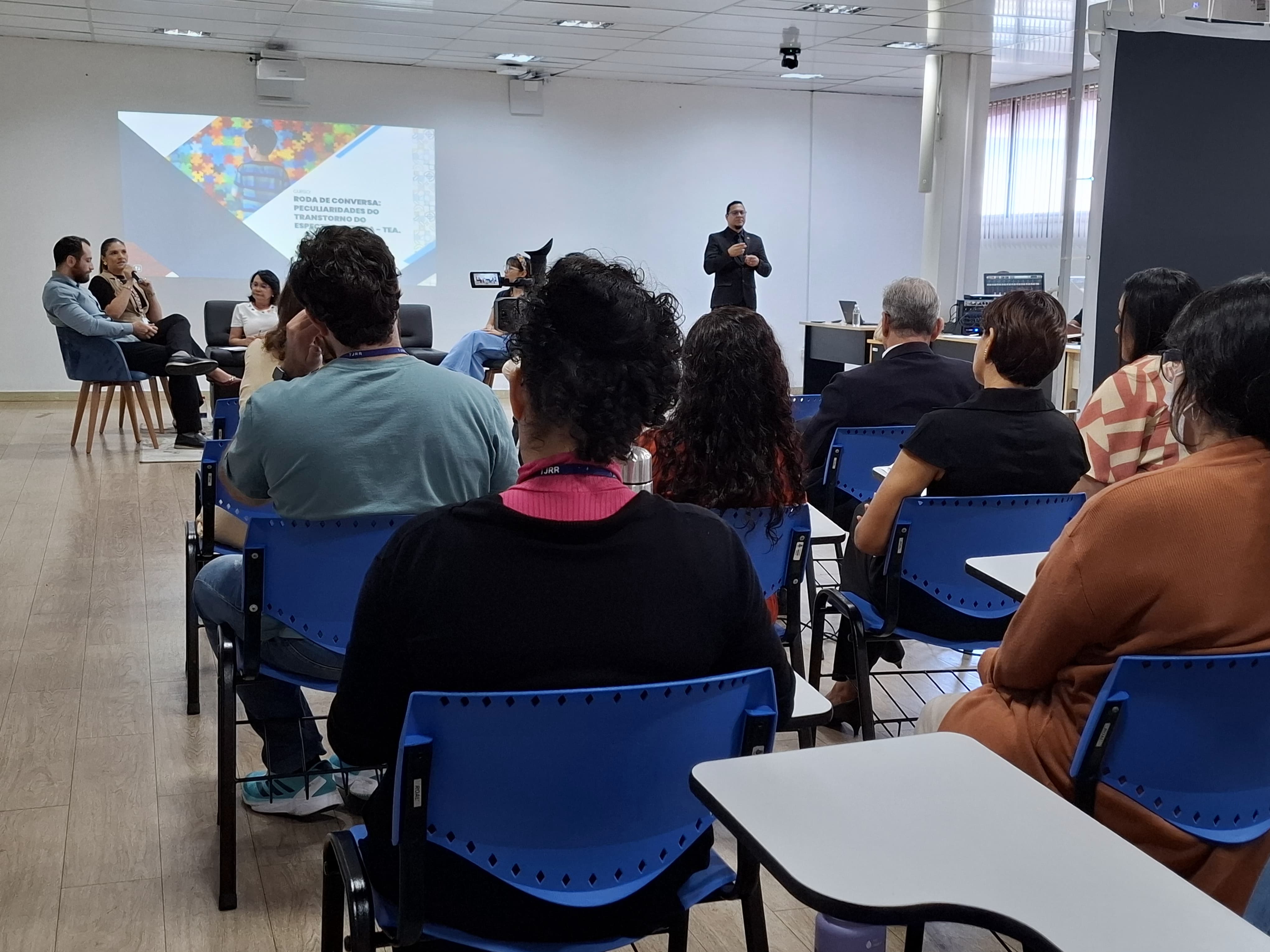 Imagem colorida contém um grupo de pessoas sentadas em cadeiras azuis, de costas para a câmera, acompanhando uma roda de conversa. À esquerda, há um telão com o título "Roda de conversa: Peculiaridades do Transtorno do Espectro Autista – TEA", com o logotipo da EJURR. Ao fundo, um intérprete de Libras está de pé, realizando a tradução simultânea.