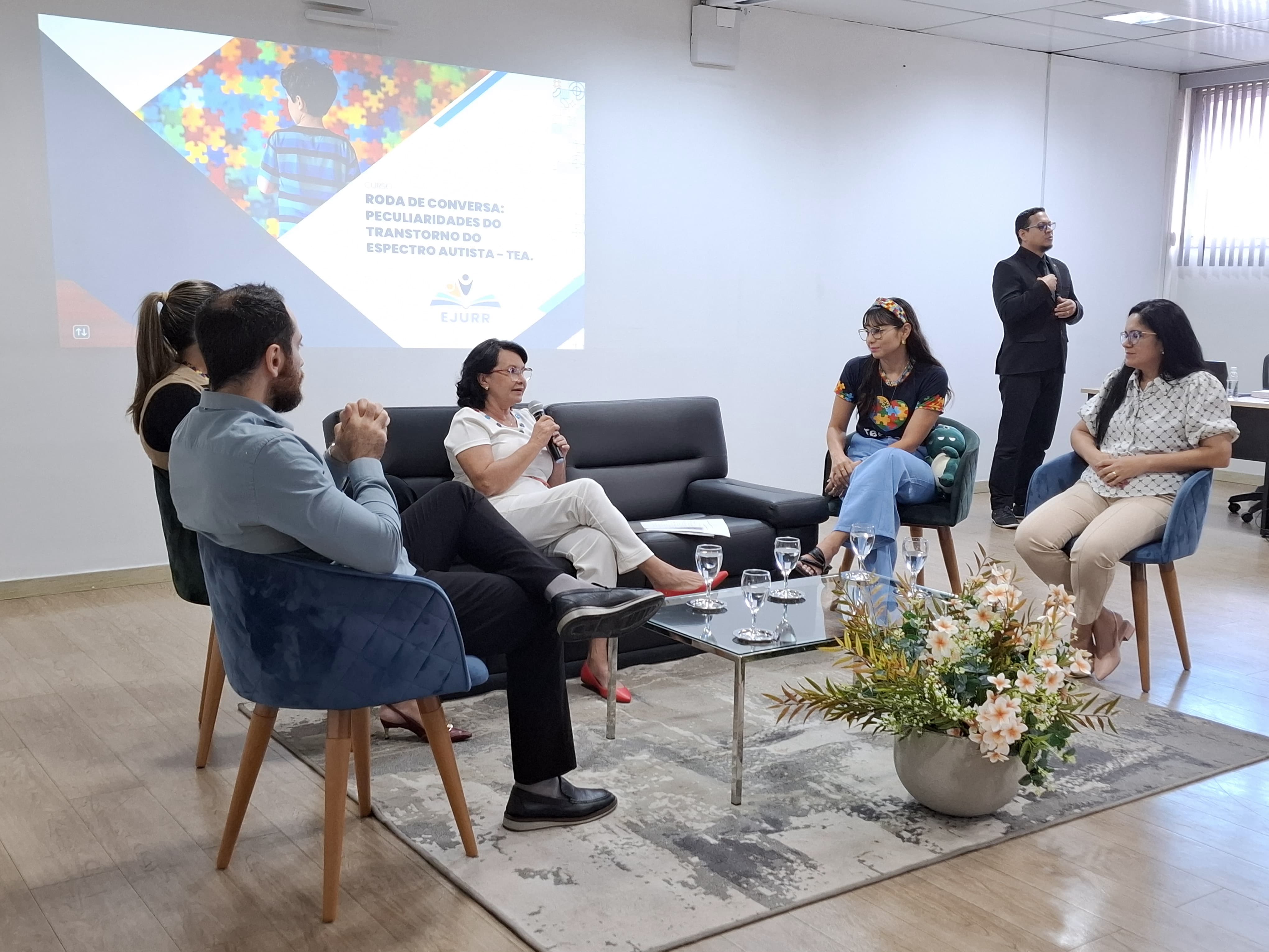 Imagem colorida contém cinco pessoas sentadas em semicírculo durante uma roda de conversa. Da esquerda para a direita: Dr. Hassan Shyaga, a psicóloga Magaly Sarah Rocha, a desembargadora Tânia Vasconcelos, a psicóloga Rozianne Melville Messa e a servidora Tatiene Quintã. A desembargadora está ao centro, sentada em um sofá preto, com papéis ao seu lado. Ela está discursando com um microfone na mão. Os demais estão em poltronas, em cima de um tapete e ao redor de uma mesa de vidro contendo taças de água. Em primeiro plano, há um arranjo de flores. Ao fundo, um telão com o título "Roda de conversa: Peculiaridades do Transtorno do Espectro Autista – TEA" e o logotipo da EJURR. À direita, um intérprete de Libras realiza a tradução simultânea.