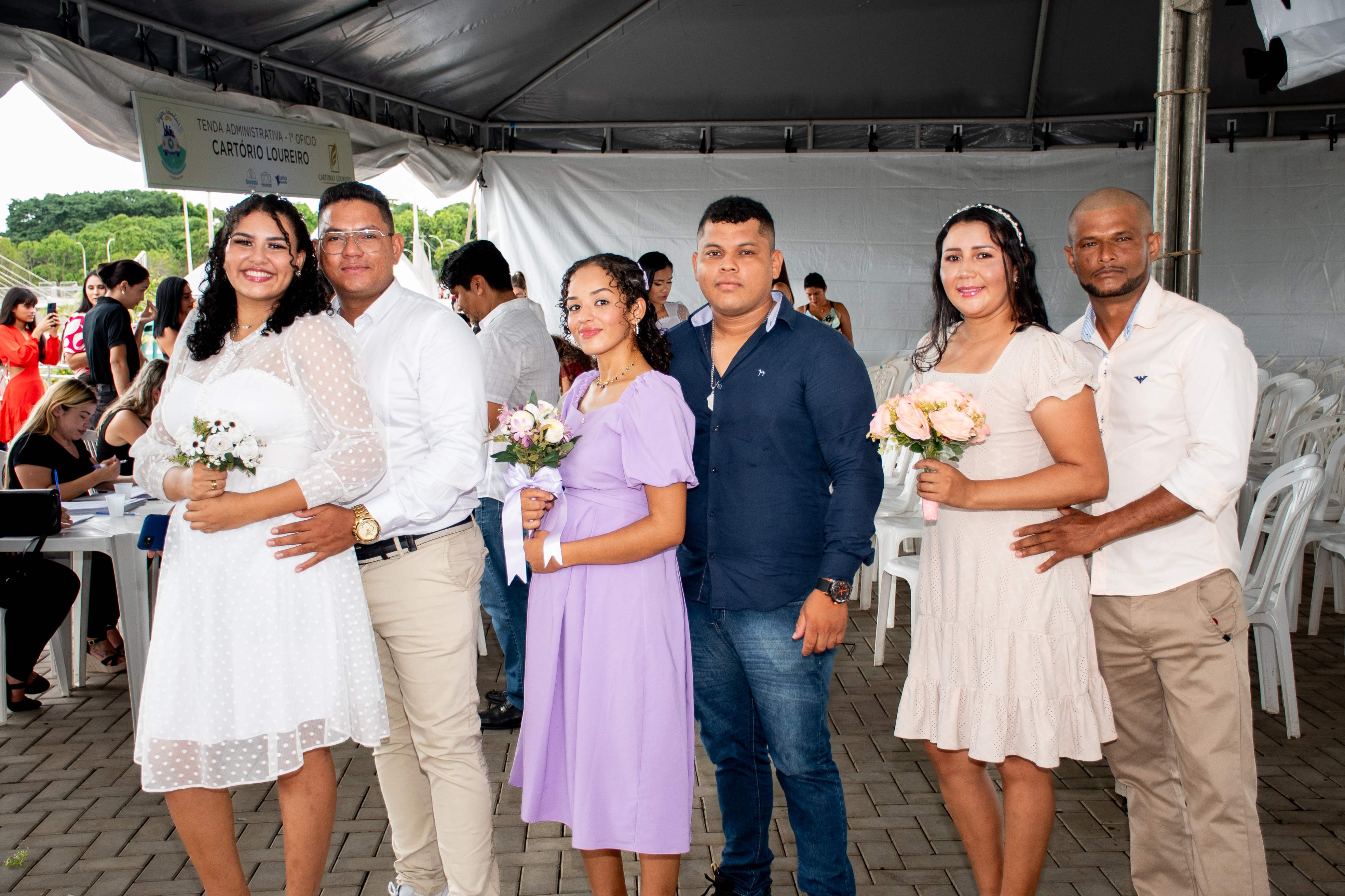 imagem colorida contém três casais, posando para fotografia, durante cerimônia matrimonial coletiva. Atrás deles encontram-se mais participantes da cerimônia.