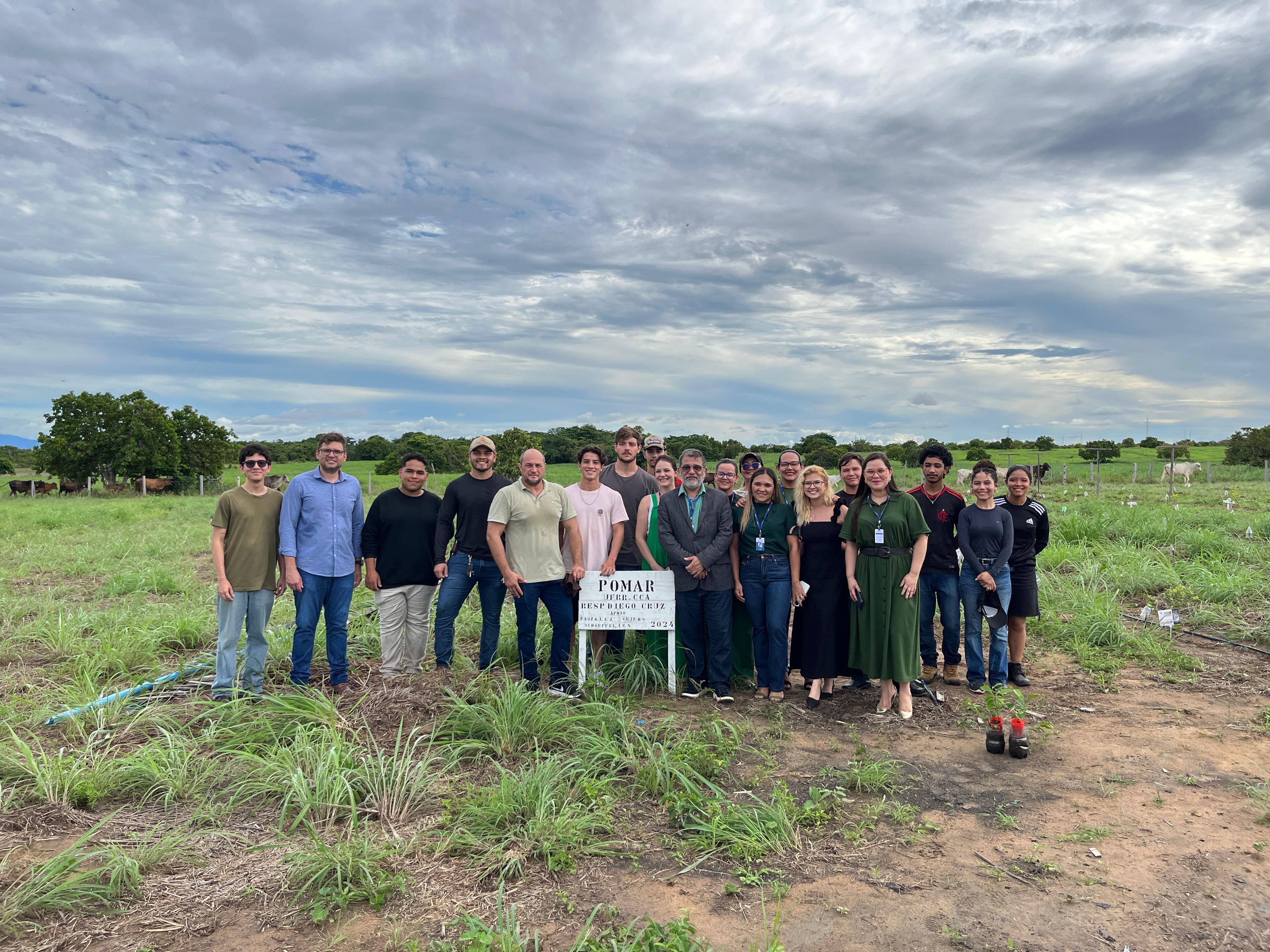 Um grupo de cerca de 20 pessoas posa para foto em frente a uma placa com informações sobre um pomar e reflorestamento, datada de agosto de 2023. A imagem, feita em um campo gramado com vegetação e animais ao fundo, indica um evento ligado a um projeto ambiental. O céu está nublado. Um grupo de cerca de 20 pessoas posa para foto em frente a uma placa com informações sobre um pomar e reflorestamento, datada de agosto de 2023. A imagem, feita em um campo gramado com vegetação e animais ao fundo, indica um evento ligado a um projeto ambiental. O céu está nublado.