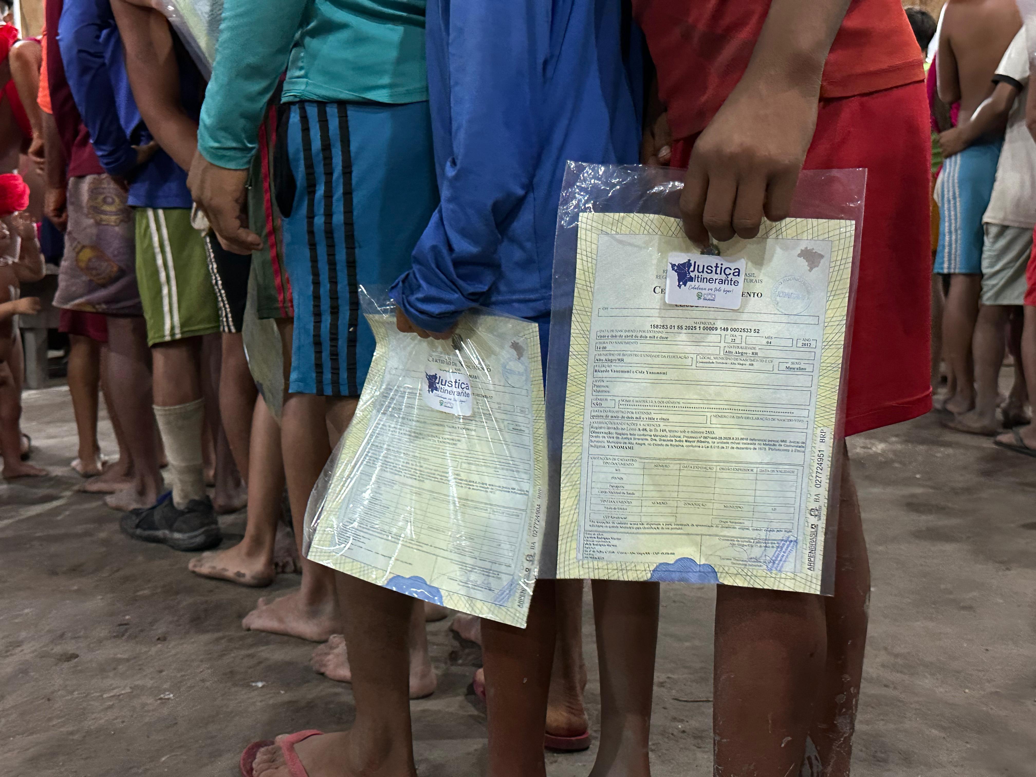 Imagem colorida contém duas pessoas em uma fila, cada uma segurando uma certidão que está em embalagem transparente com o selo da Justiça Itinerante.