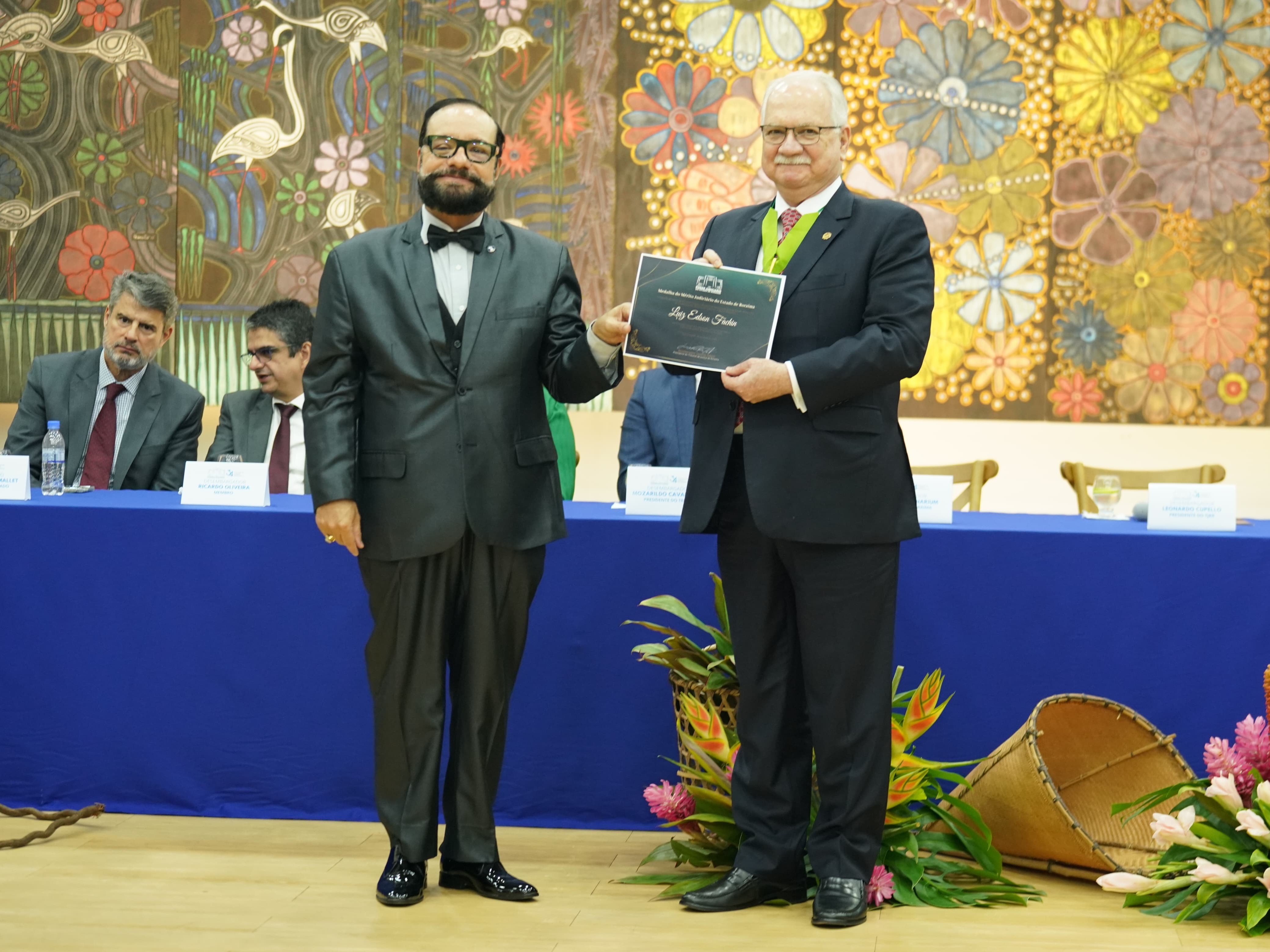 Foto colorida mostra o presidente do TJRR, desembargador Leonardo Cupello, posando para fotografia ao lado do ministro do STF, Edson Fachin. O ministro segura o certificado da Medalha do Mérito Judiciário do Estado de Roraima. Ao fundo, vê-se a mesa de autoridades. 