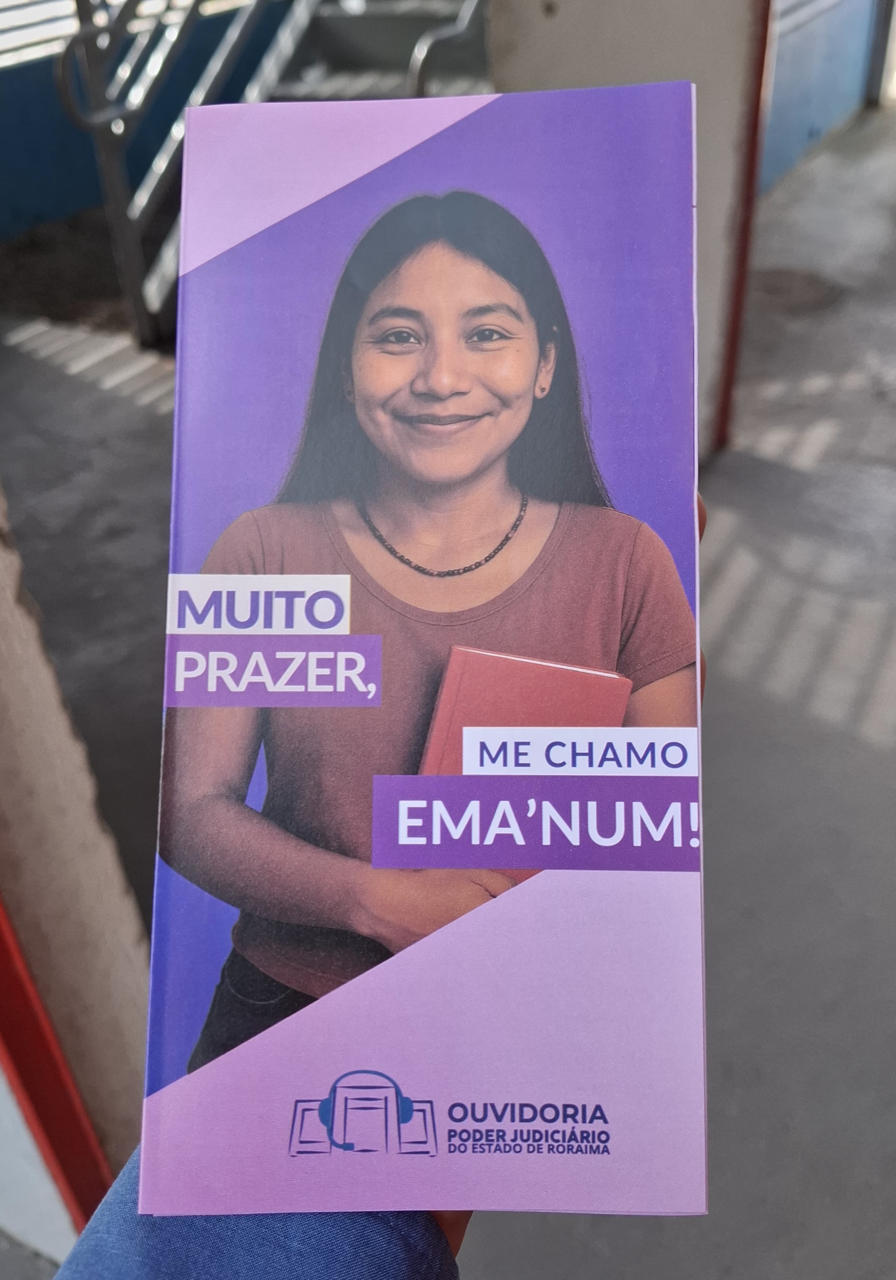 Uma pessoa segura um panfleto vertical em suas mãos. O panfleto tem um fundo lilás e apresenta a foto de uma jovem com cabelos longos e escuros, vestindo uma camiseta marrom e segurando um livro vermelho. Ao lado da foto, em letras brancas, está escrito "MUITO PRAZER," à esquerda e "ME CHAMO EMA'NUM!" à direita. Na parte inferior do panfleto, há um logotipo branco com a inscrição "OUVIDORIA" acima de "PODER JUDICIÁRIO DO ESTADO DE RORAIMA". Uma pessoa segura um panfleto vertical em suas mãos. O panfleto tem um fundo lilás e apresenta a foto de uma jovem com cabelos longos e escuros, vestindo uma camiseta marrom e segurando um livro vermelho. Ao lado da foto, em letras brancas, está escrito "MUITO PRAZER," à esquerda e "ME CHAMO EMA'NUM!" à direita. Na parte inferior do panfleto, há um logotipo branco com a inscrição "OUVIDORIA" acima de "PODER JUDICIÁRIO DO ESTADO DE RORAIMA".