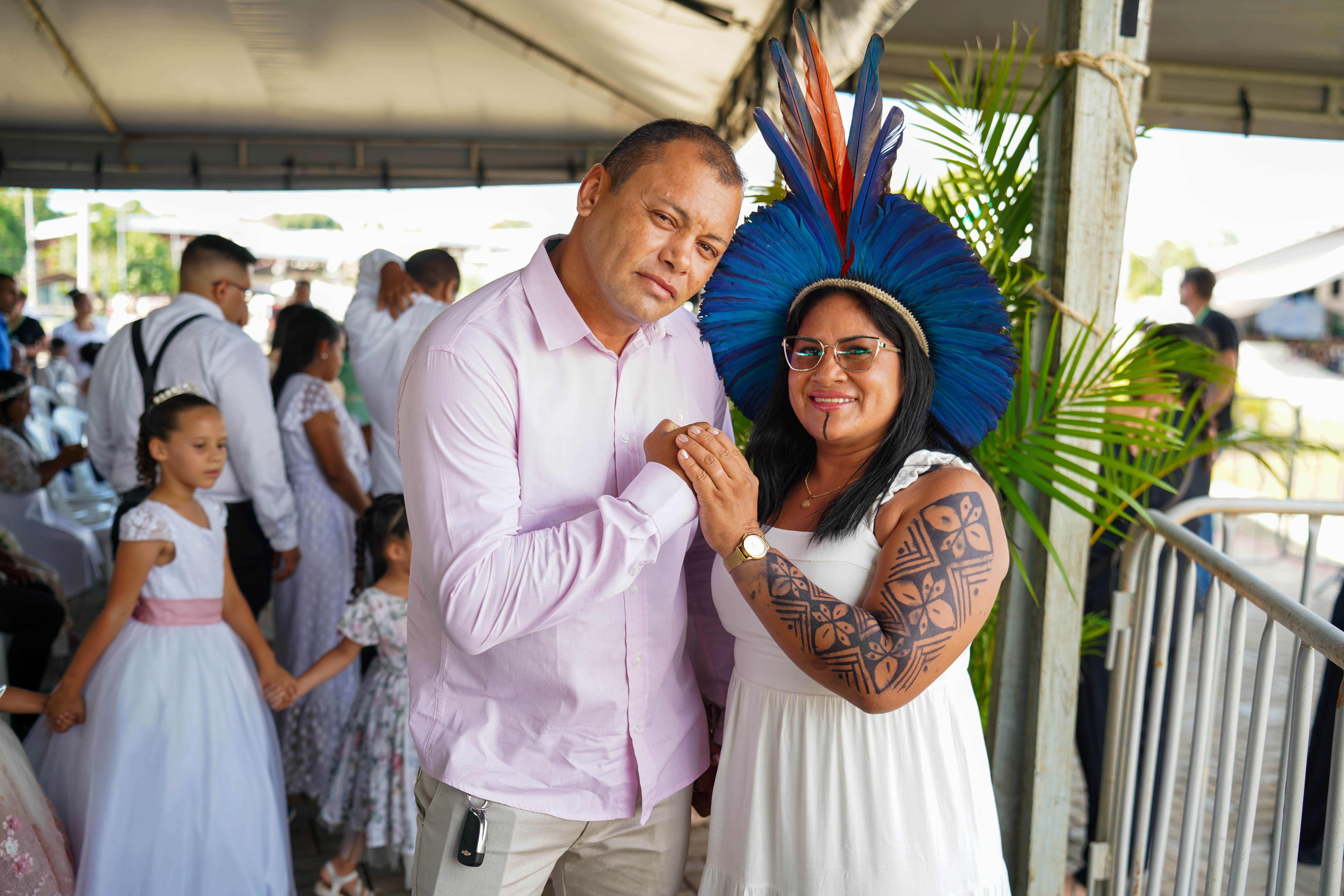 Foto colorida mostra um casal em primeiro plano, posando para a câmera. O homem, à esquerda, usa uma camisa social rosa clara. A mulher, à direita, usa um vestido branco e um cocar vibrante de penas azuis e laranjas. Ela tem tatuagens com grafismos indígenas em seu braço direito. Ao fundo, outras pessoas e parte de uma estrutura de tenda são visíveis. Foto colorida mostra um casal em primeiro plano, posando para a câmera. O homem, à esquerda, usa uma camisa social rosa clara. A mulher, à direita, usa um vestido branco e um cocar vibrante de penas azuis e laranjas. Ela tem tatuagens com grafismos indígenas em seu braço direito. Ao fundo, outras pessoas e parte de uma estrutura de tenda são visíveis.