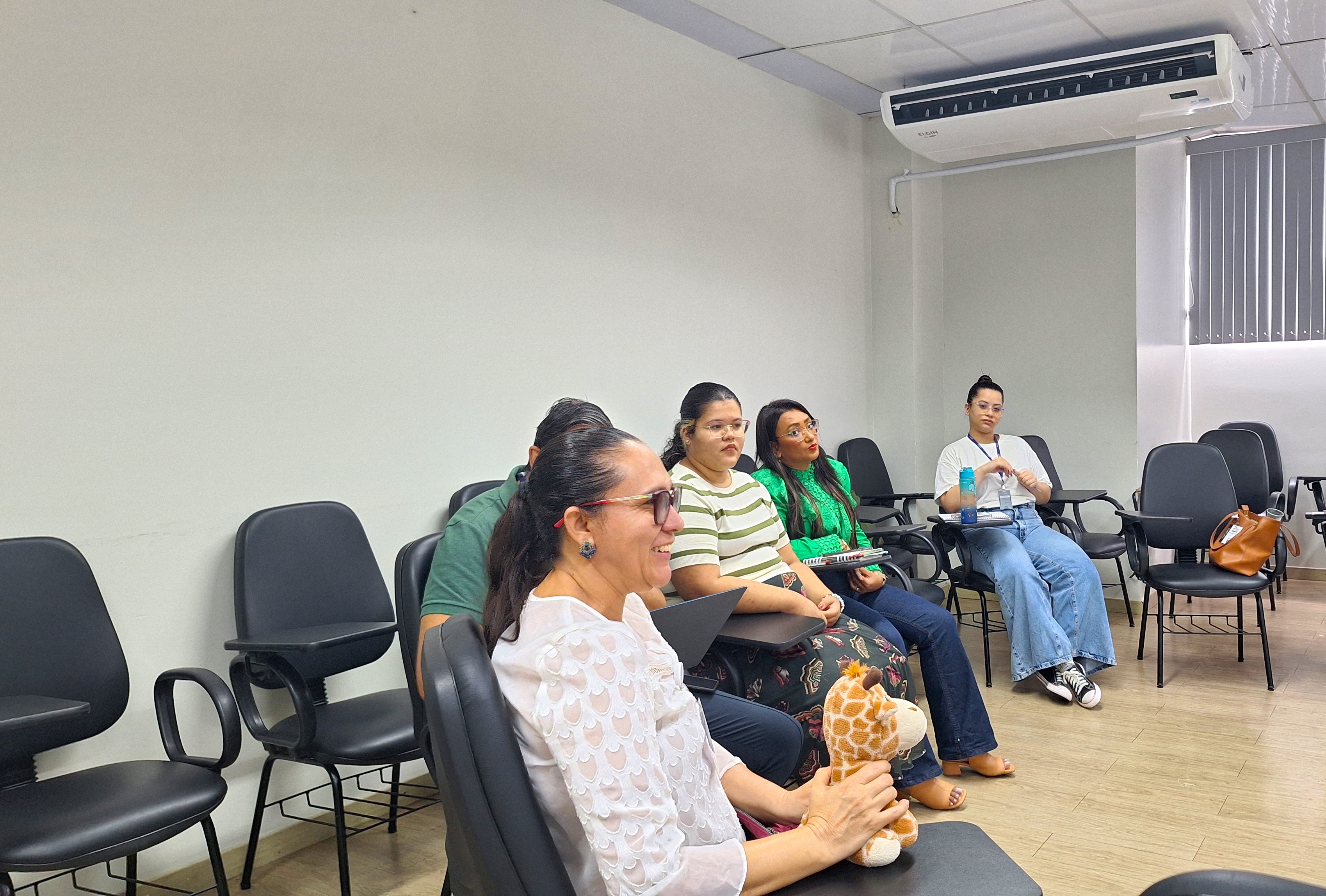 foto colorida dando enfoque no meio circulo de participantes. Em primeiro plano está uma participante sorrindo enquanto segura uma girafa de pelúcia. foto colorida dando enfoque no meio circulo de participantes. Em primeiro plano está uma participante sorrindo enquanto segura uma girafa de pelúcia.