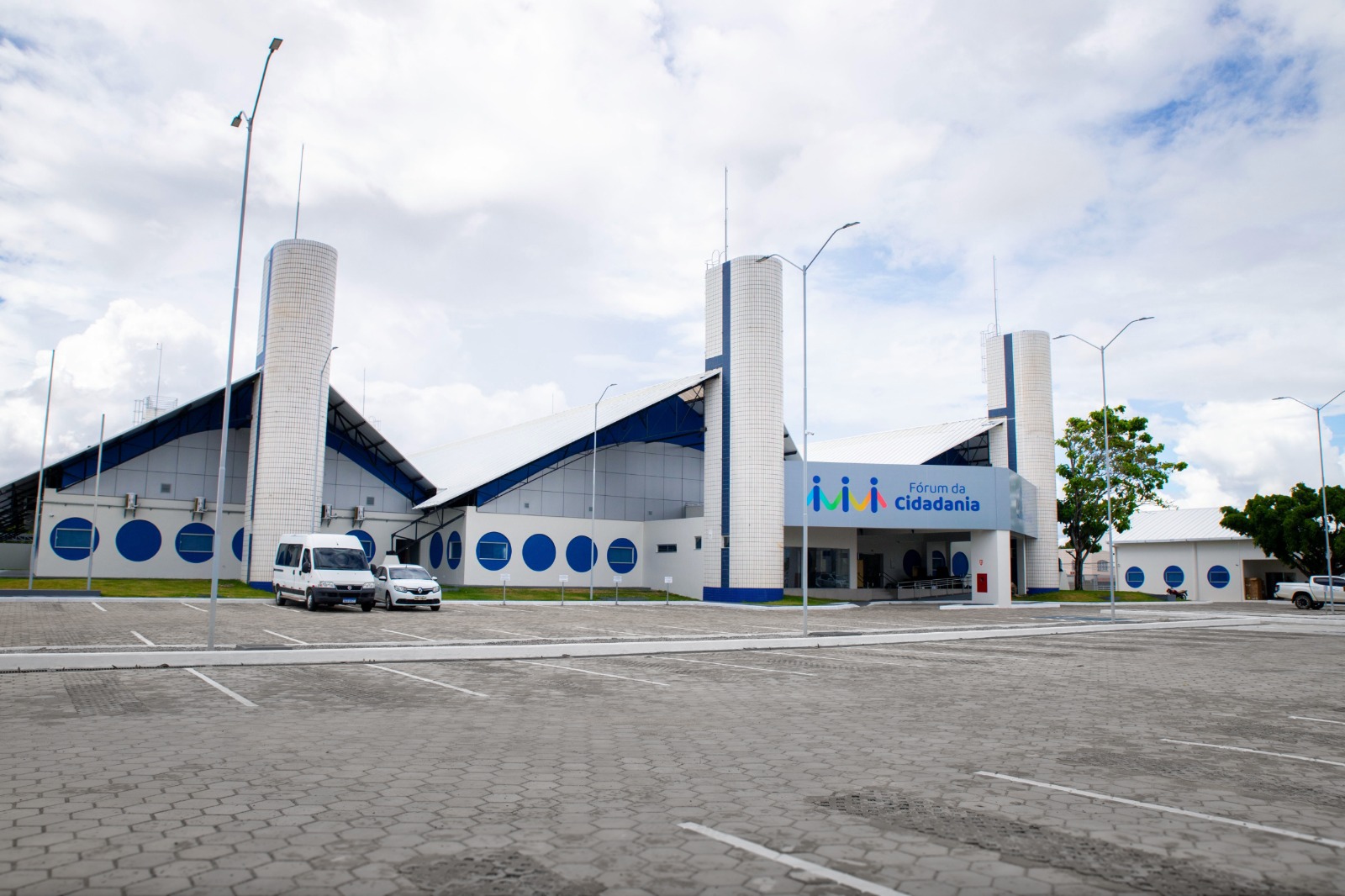 Foto colorida mostra a fachada do Fórum da Cidadania. O prédio possui design geométrico e janelas redondas azuis. A entrada tem um letreiro com o nome e um logotipo colorido. Há poucos carros no estacionamento amplo e o local é bem estruturado, com rampa de acessibilidade Foto colorida mostra a fachada do Fórum da Cidadania. O prédio possui design geométrico e janelas redondas azuis. A entrada tem um letreiro com o nome e um logotipo colorido. Há poucos carros no estacionamento amplo e o local é bem estruturado, com rampa de acessibilidade