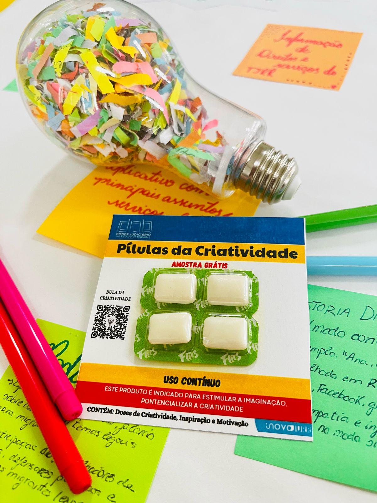 imagem colorida de uma mesa com diversos objetos relacionados à criatividade e ideias. No centro, destaca-se uma embalagem que se assemelha a um blister de remédios, intitulada "Pílulas da Criatividade". A embalagem indica ser uma "AMOSTRA GRÁTIS" e descreve o produto como sendo para "USO CONTÍNUO". Ao lado da embalagem, há uma lâmpada de vidro transparente preenchida com pequenos pedaços coloridos de papel, sugerindo a metáfora de uma "explosão" de ideias. imagem colorida de uma mesa com diversos objetos relacionados à criatividade e ideias. No centro, destaca-se uma embalagem que se assemelha a um blister de remédios, intitulada "Pílulas da Criatividade". A embalagem indica ser uma "AMOSTRA GRÁTIS" e descreve o produto como sendo para "USO CONTÍNUO". Ao lado da embalagem, há uma lâmpada de vidro transparente preenchida com pequenos pedaços coloridos de papel, sugerindo a metáfora de uma "explosão" de ideias.