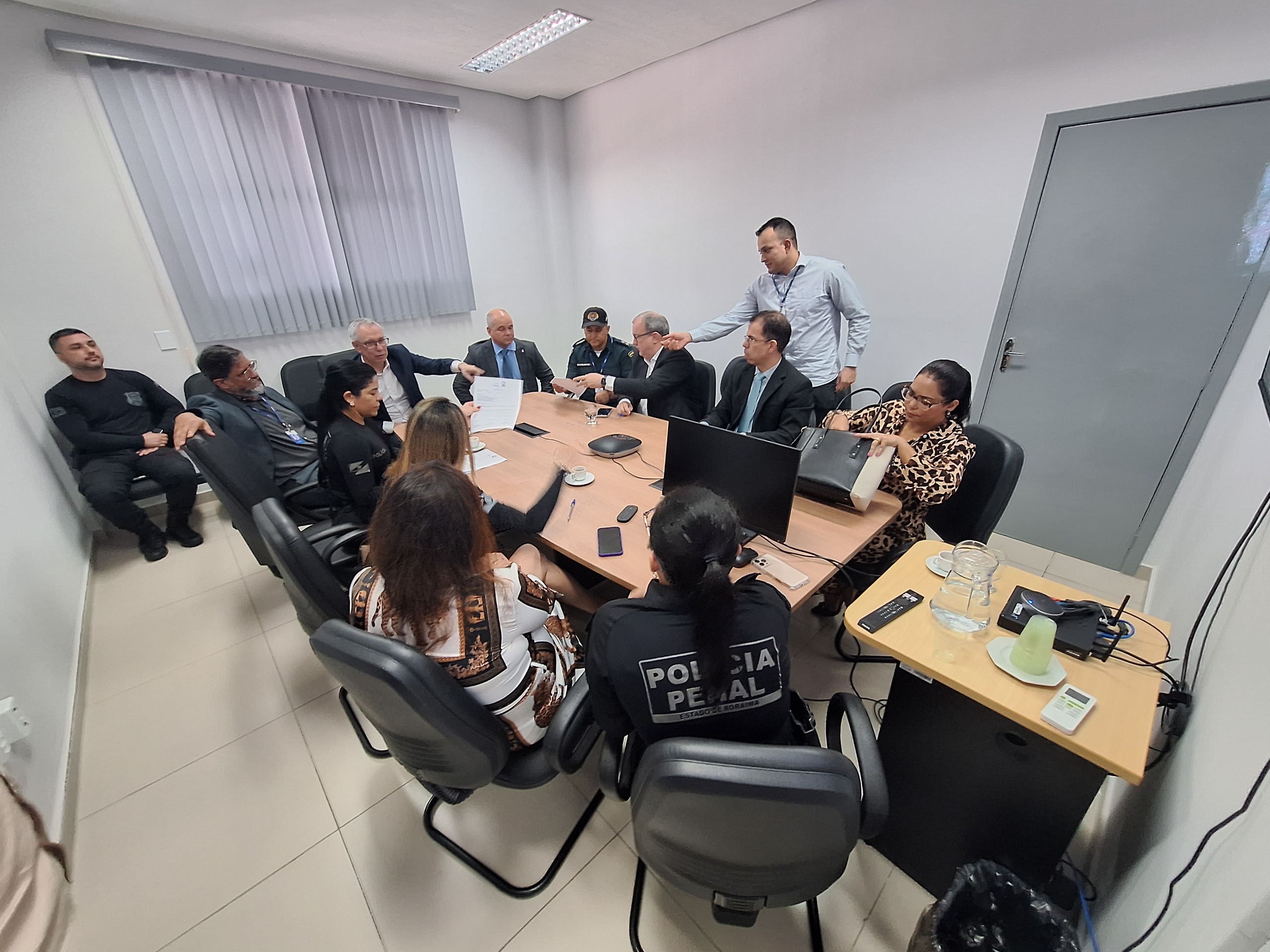 Foto colorida mostra uma reunião formal em andamento, realizada em uma sala fechada. Ao redor de uma mesa retangular estão sentadas cerca de treze pessoas, entre elas homens e mulheres, com diferentes trajes — alguns em ternos, outros com uniformes da Polícia Penal.