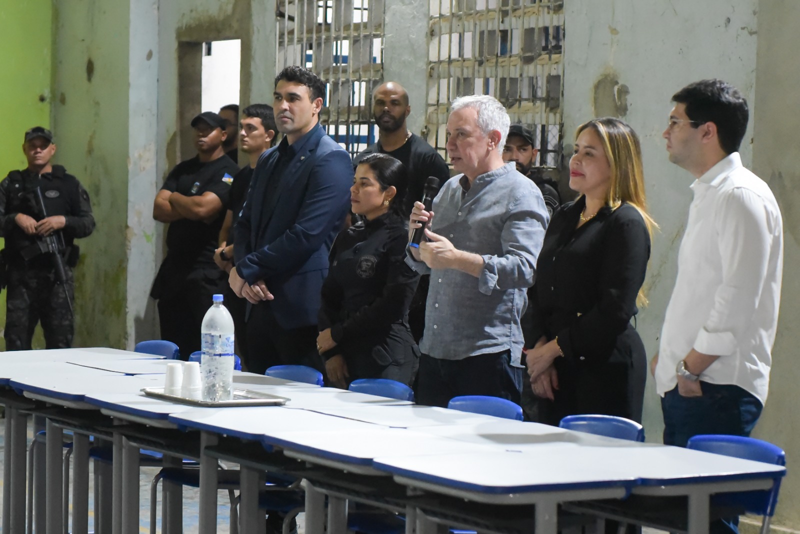 Imagem colorida mostra um grupo de pessoas em pé, incluindo homens e mulheres, alguns em roupas sociais escuras e claras, e outros em uniformes. Ao centro, um homem decabelo grisalho com uma camisa social cinza clara está segurando um microfone e falando. À direita, há uma mesa comprida com garrafas de água e copos. Ao fundo, uma grade na janela.