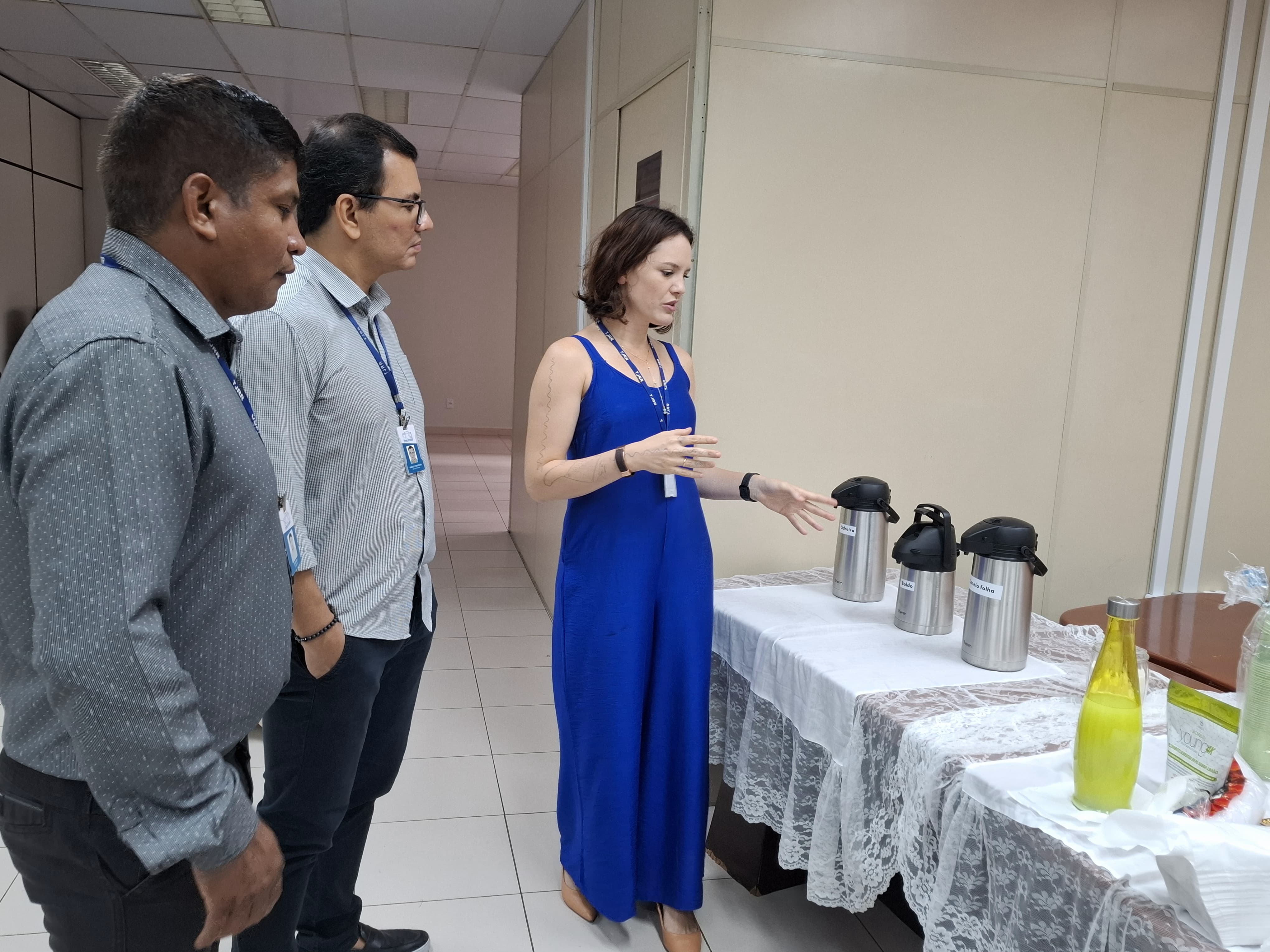 Imagem colorida mostra três pessoas em um corredor ou sala. Dois homens estão à esquerda, olhando para uma mulher que está à direita. Ela está gesticulando em direção a uma mesa coberta por uma toalha branca de renda, onde estão dispostas garrafas térmicas prateadas e uma garrafa de líquido amarelo. Os homens parecem estar ouvindo atentamente a explicação da mulher.