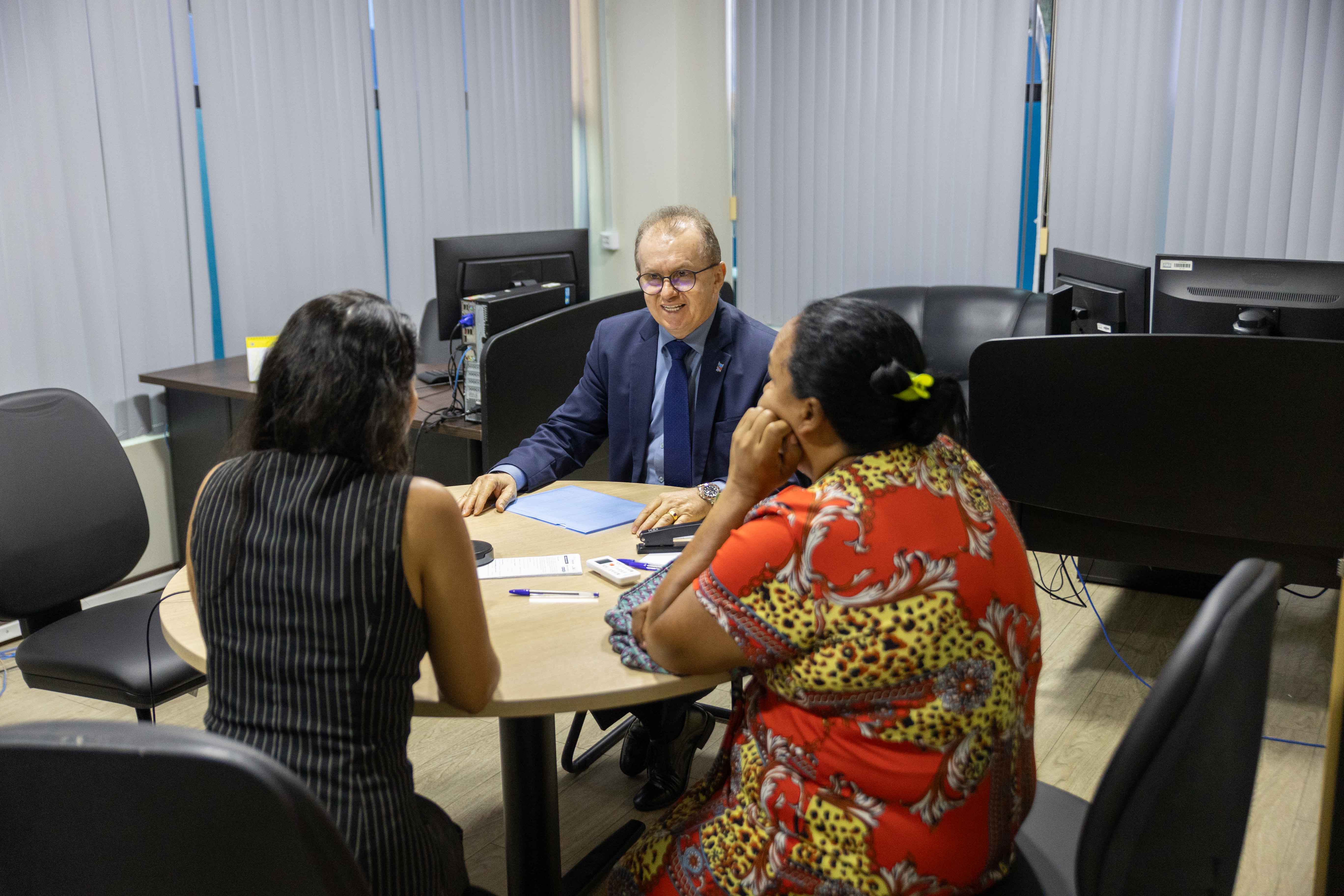 Foto colorida mostra três pessoas sentadas a mesa conversando. Um homem vestido com terno azul escuro e gravata, conversa com duas mulheres em uma sala com ambiente formal. Todos estão sentados em torno de uma mesa redonda, participando de uma audiência de conciliação.