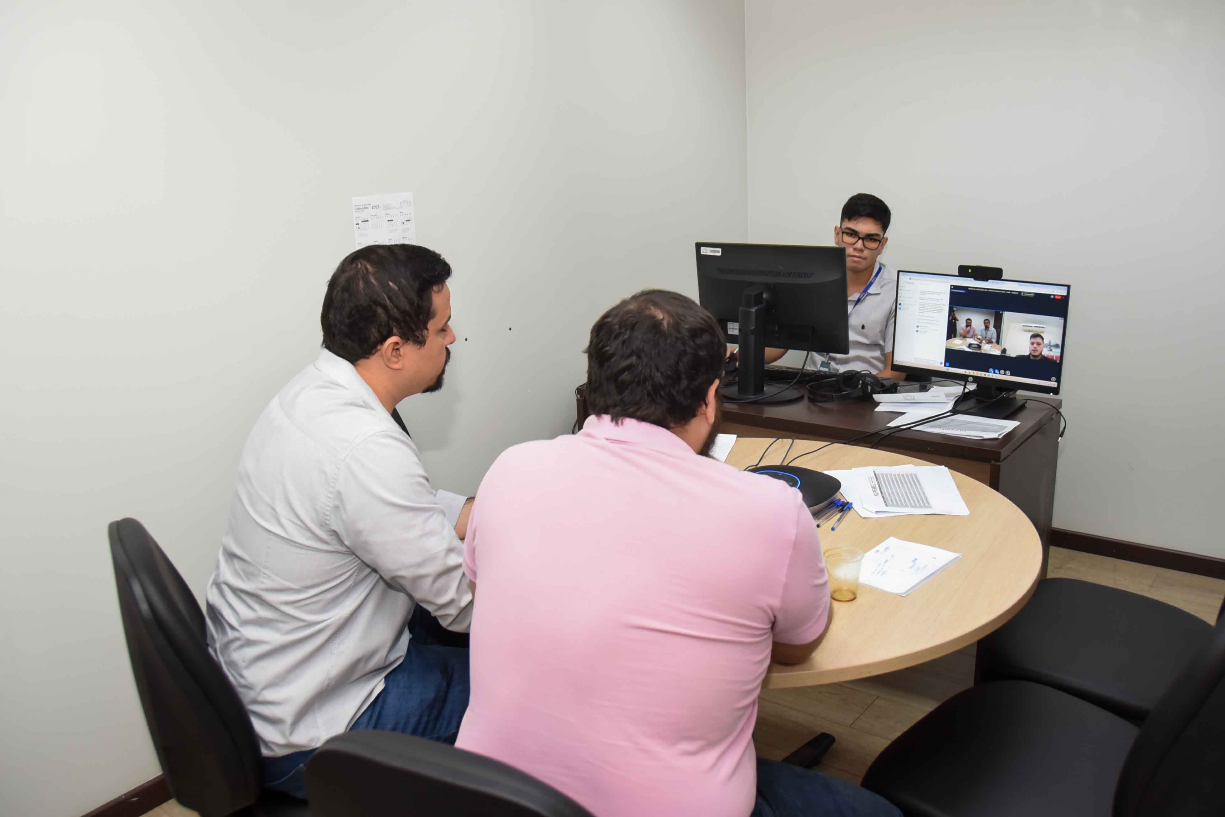 Foto colorida mostra três homens participando de uma audiência de mediação híbrida. Dois estão sentados presencialmente em frente a um servidor que conduz a sessão, enquanto outras pessoas aparecem em uma chamada de vídeo na tela do computador.