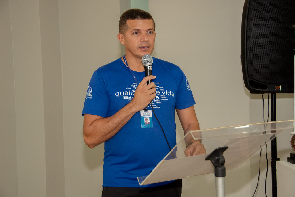 Um servidor do Tribunal de Justiça de Roraima, vestido com a camiseta azul do evento, fala ao microfone em um púlpito acrílico durante a cerimônia de encerramento do desafio. Ele usa crachá funcional e tem ao fundo uma caixa de som e parte da decoração do ambiente. Um servidor do Tribunal de Justiça de Roraima, vestido com a camiseta azul do evento, fala ao microfone em um púlpito acrílico durante a cerimônia de encerramento do desafio. Ele usa crachá funcional e tem ao fundo uma caixa de som e parte da decoração do ambiente.