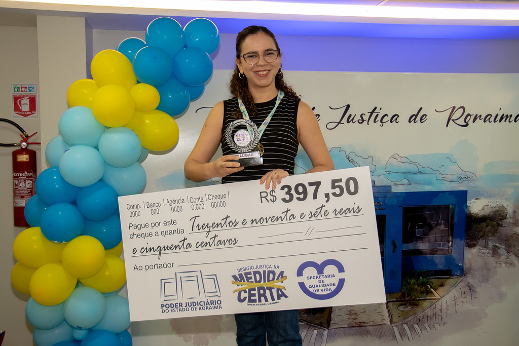 Uma mulher posa orgulhosa segurando o troféu e um cheque simbólico no valor de R$ 397,50. Ela está à frente do painel institucional com o nome do Tribunal de Justiça de Roraima, e ao lado de uma decoração com balões azuis e amarelos. Usa óculos, blusa preta com listras e calça jeans. Uma mulher posa orgulhosa segurando o troféu e um cheque simbólico no valor de R$ 397,50. Ela está à frente do painel institucional com o nome do Tribunal de Justiça de Roraima, e ao lado de uma decoração com balões azuis e amarelos. Usa óculos, blusa preta com listras e calça jeans.