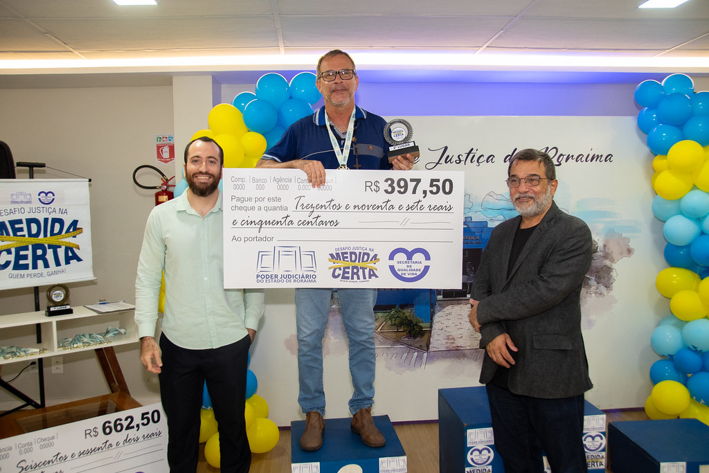 Três homens posam sorridentes para a foto durante a premiação do Desafio Justiça na Medida Certa. O homem ao centro, em um pódio, segura um troféu e um cheque simbólico no valor de R$ 397,50. Ele veste uma camisa azul e jeans. Ao fundo, há balões nas cores azul, amarelo e branco, e o painel com os dizeres "Justiça de Roraima". Três homens posam sorridentes para a foto durante a premiação do Desafio Justiça na Medida Certa. O homem ao centro, em um pódio, segura um troféu e um cheque simbólico no valor de R$ 397,50. Ele veste uma camisa azul e jeans. Ao fundo, há balões nas cores azul, amarelo e branco, e o painel com os dizeres "Justiça de Roraima".