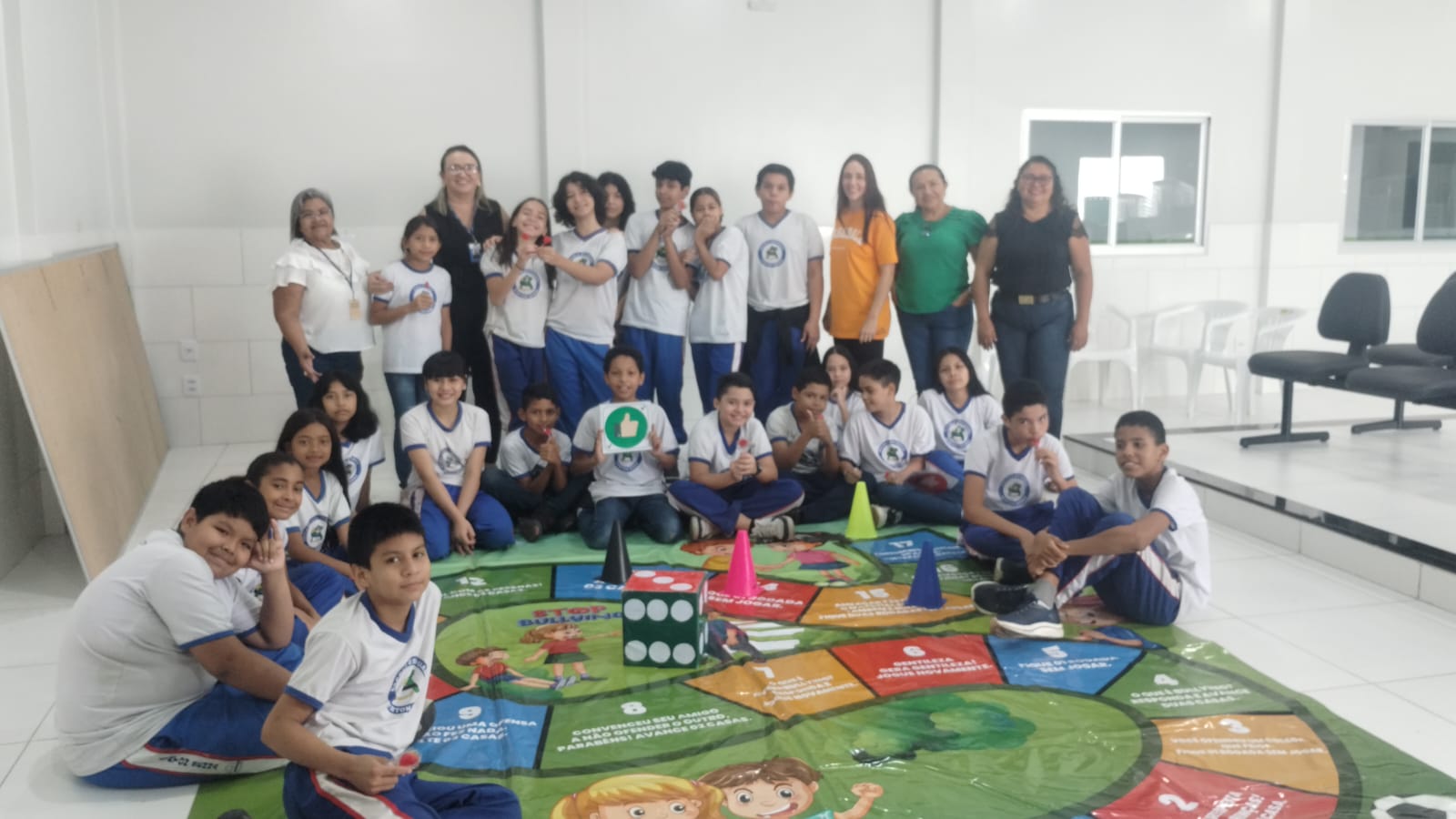 A imagem colorida mostra um grupo de cerca de 15 pessoas, incluindo adultos e jovens, participando de uma atividade em um espaço coberto. A maioria dos jovens veste uniformes escolares, e alguns adultos parecem ser educadores. As pessoas estão em círculo, algumas com os braços estendidos e segurando canetas, sugerindo um jogo ou exercício de equipe. Há uma parede clara com um padrão geométrico decorativo e, ao fundo, árvores e um carro. A imagem colorida mostra um grupo de cerca de 15 pessoas, incluindo adultos e jovens, participando de uma atividade em um espaço coberto. A maioria dos jovens veste uniformes escolares, e alguns adultos parecem ser educadores. As pessoas estão em círculo, algumas com os braços estendidos e segurando canetas, sugerindo um jogo ou exercício de equipe. Há uma parede clara com um padrão geométrico decorativo e, ao fundo, árvores e um carro.