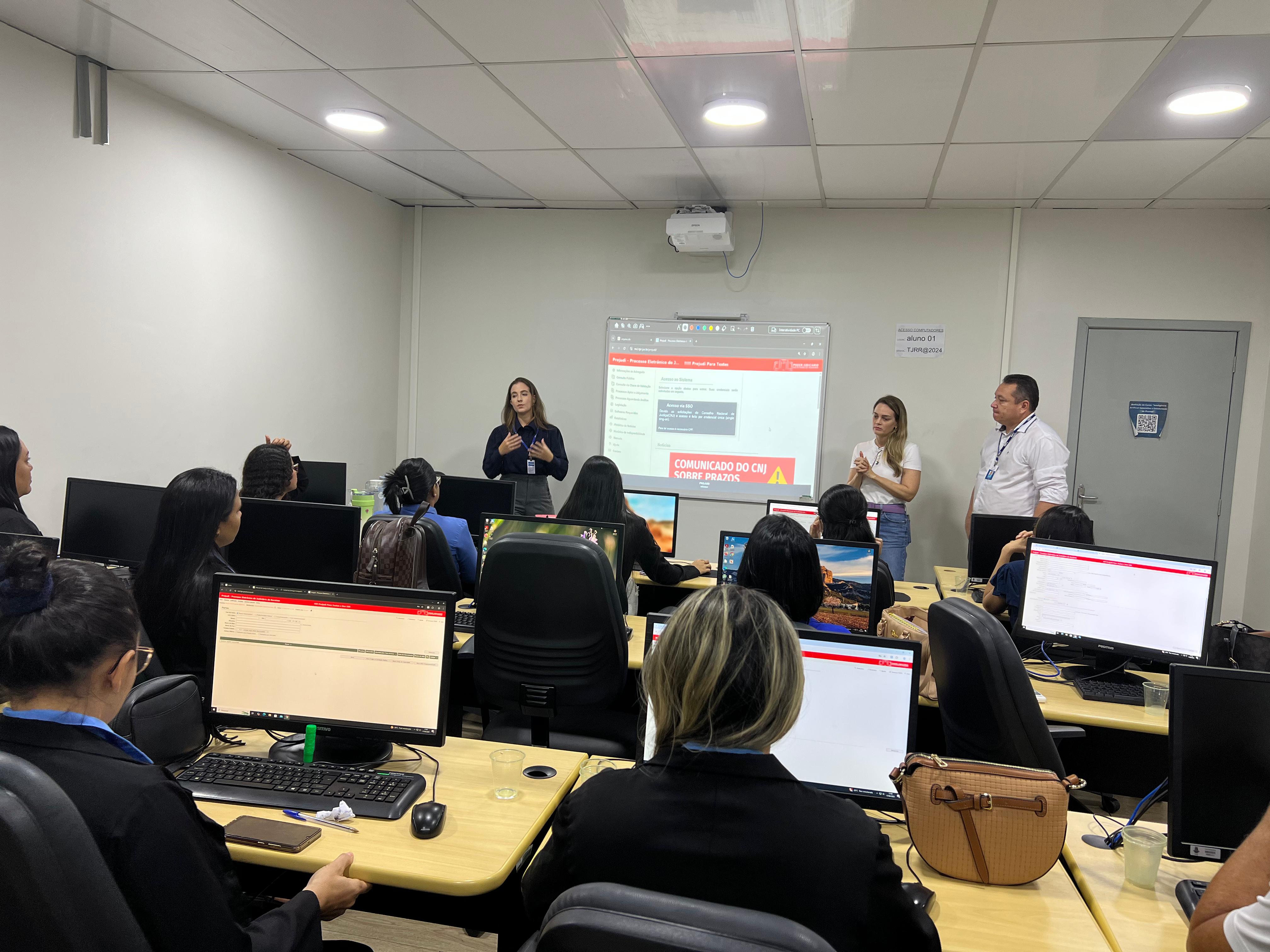  Foto colorida mostra várias pessoas em uma sala de informática participando de uma capacitação. Três instrutores estão em pé à frente da sala — duas mulheres e um homem — enquanto uma das facilitadoras fala aos participantes. No ambiente, todos os alunos estão sentados diante de computadores, acompanhando o conteúdo projetado na parede, que exibe uma página com o título “COMUNICADO DO CNJ SOBRE PRAZOS”. 