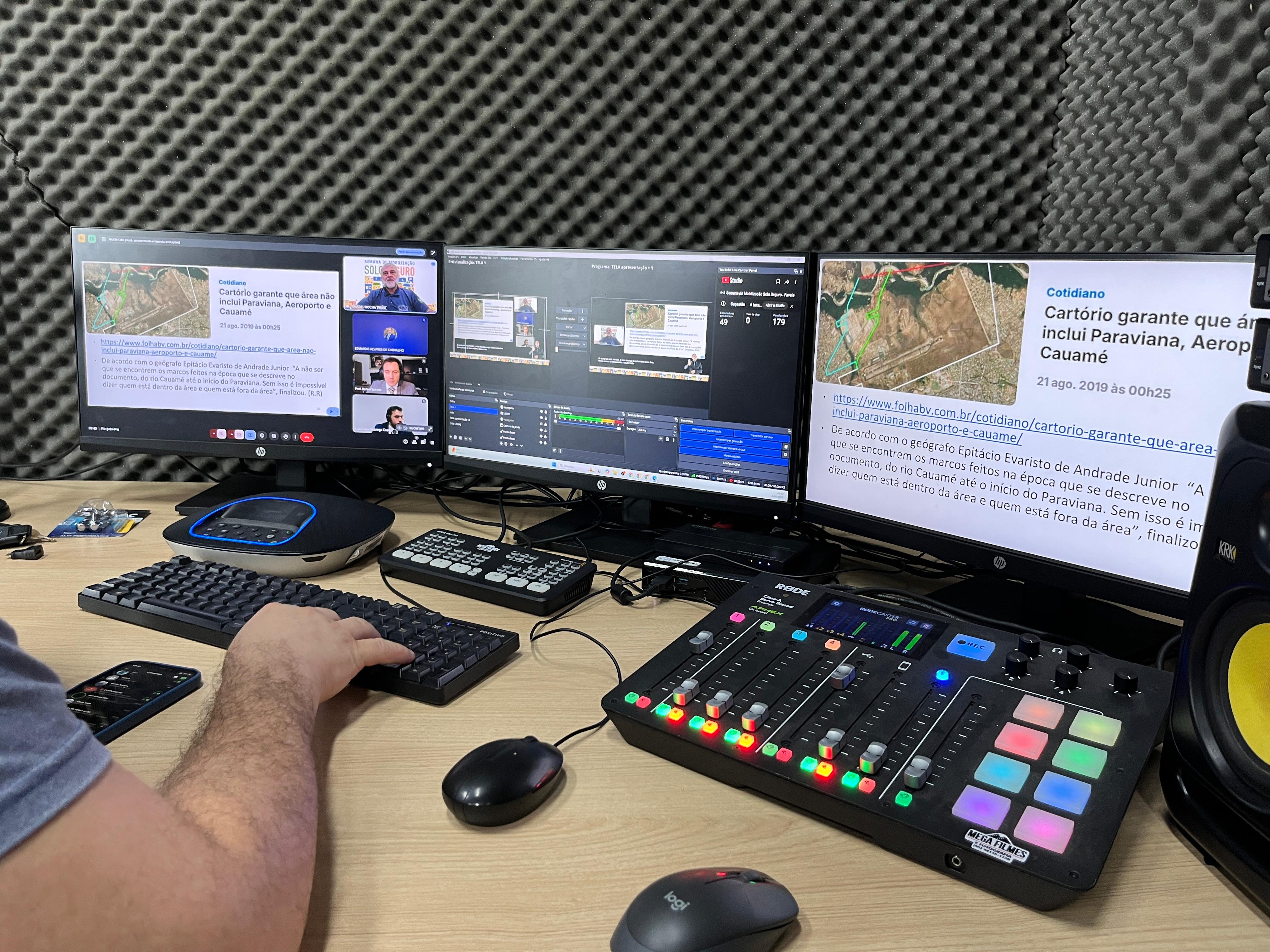 Foto colorida mostra um operador de audiovisual trabalhando em uma bancada equipada com três monitores, um teclado, uma mesa de som e outros equipamentos de transmissão. As telas exibem uma transmissão ao vivo com apresentações e reportagens relacionadas à regularização fundiária. O ambiente é tratado acusticamente com placas de isolamento sonoro nas paredes Foto colorida mostra um operador de audiovisual trabalhando em uma bancada equipada com três monitores, um teclado, uma mesa de som e outros equipamentos de transmissão. As telas exibem uma transmissão ao vivo com apresentações e reportagens relacionadas à regularização fundiária. O ambiente é tratado acusticamente com placas de isolamento sonoro nas paredes