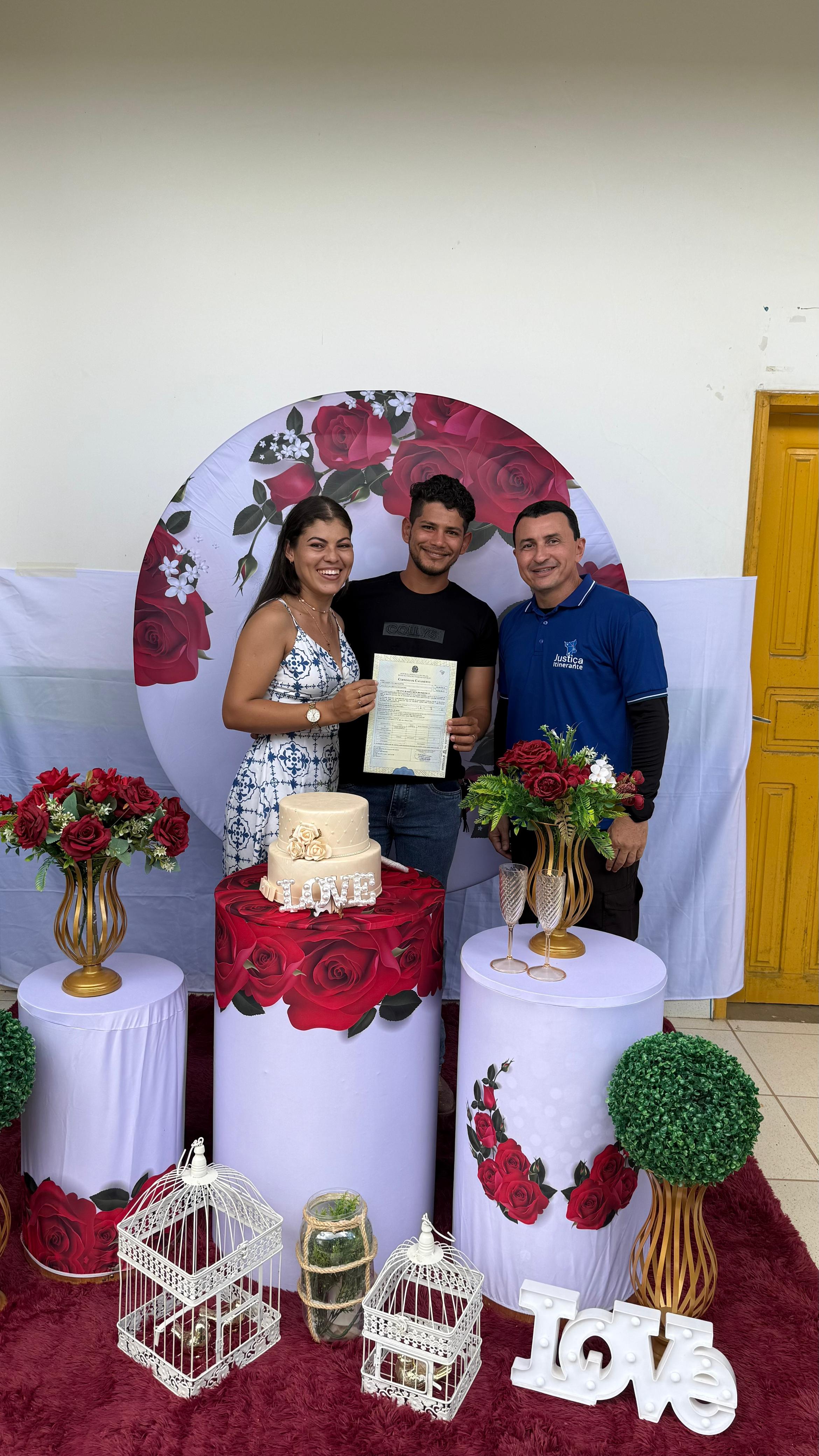 A imagem mostra um casal celebrando sua união civil durante atendimento da Vara da Justiça Itinernate. A mulher veste um vestido azul e branco, e o homem segura uma certidão de casamento, sorrindo. Ao lado deles, um servidor com camisa azul posa com a mão no ombro do noivo. Ao fundo, há uma decoração com rosas vermelhas, bolo, flores e a palavra “Love”. A cena transmite alegria e acolhimento em um ambiente festivo e simples.