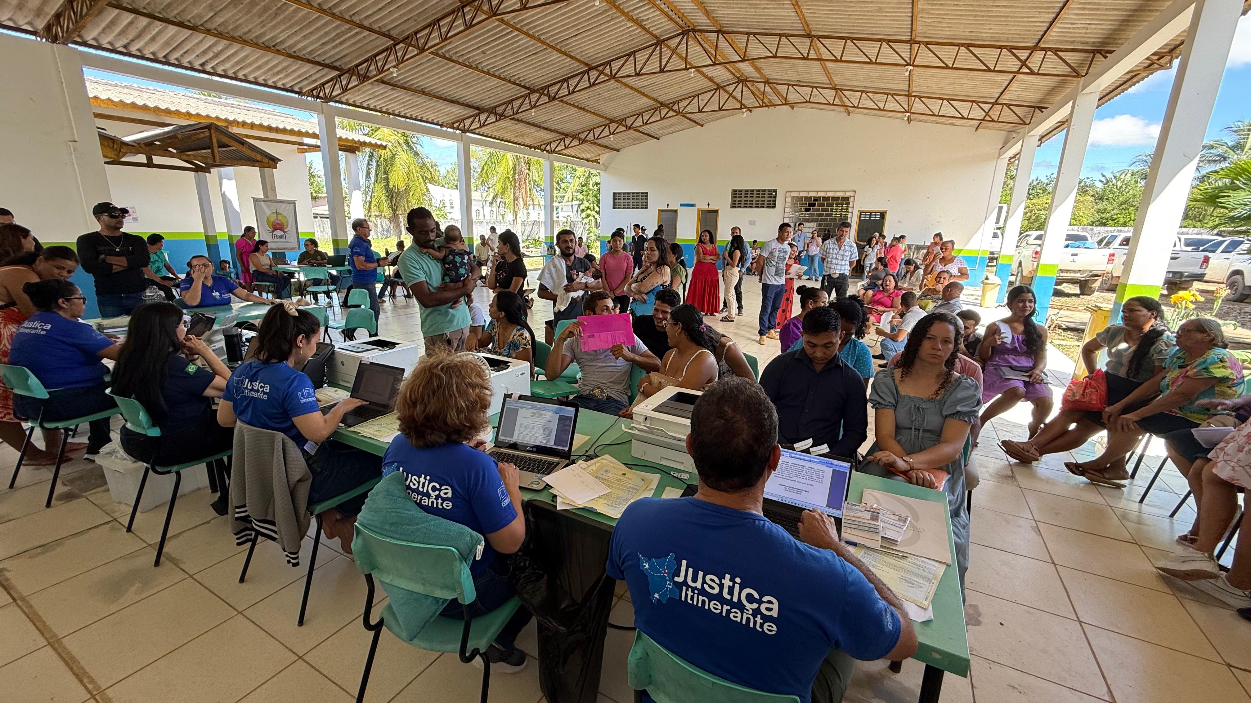 A imagem mostra um evento da Justiça Itinerante acontecendo durante o dia em um espaço aberto e coberto, com palmeiras ao fundo. Em destaque, duas pessoas de camiseta azul com a inscrição "Justiça Itinerante" atendem cidadãos em mesas com laptops. A comunidade comparece em peso, com pessoas sentadas, em filas ou conversando. O ambiente é simples, com telhado metálico e iluminação natural. A cena retrata um dia ativo de prestação de serviços jurídicos à população.