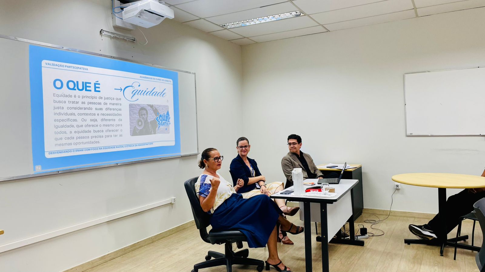 Foto 2: Imagem colorida mostra três pessoas sentadas à frente de uma sala de aula durante uma reunião com estudantes sobre equidade racial no Exame Nacional da Magistratura. Um gráfico circular com dados sobre equidade racial na magistratura brasileira é projetado no quadro branco ao fundo. Algumas pessoas assistem sentadas em cadeiras.