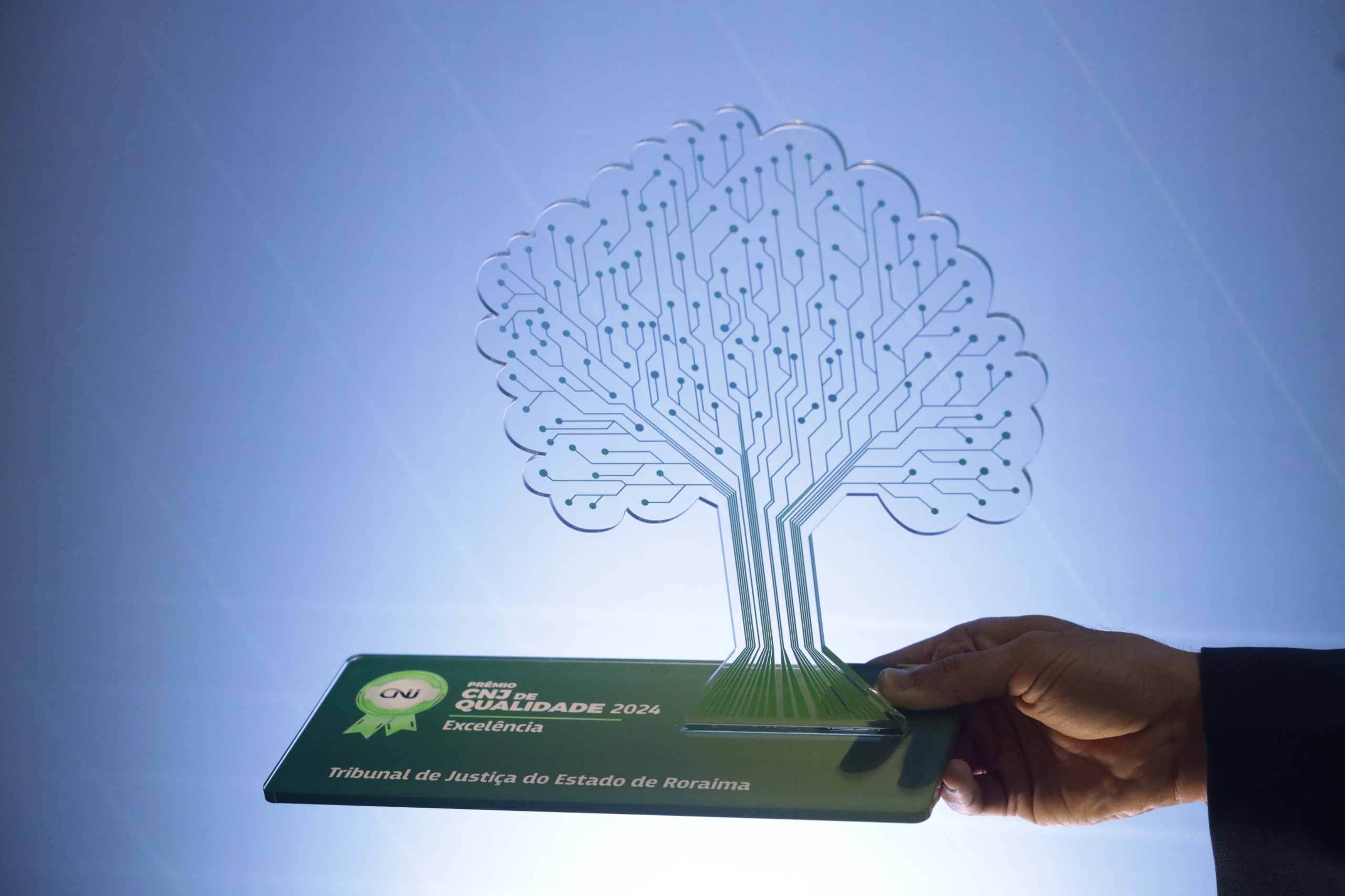 A imagem mostra um troféu transparente em formato de árvore, com linhas que lembram circuitos eletrônicos representando os galhos. A base do troféu é verde, com a inscrição "Prêmio A imagem mostra um troféu transparente em formato de árvore, com linhas que lembram circuitos eletrônicos representando os galhos. A base do troféu é verde, com a inscrição "Prêmio