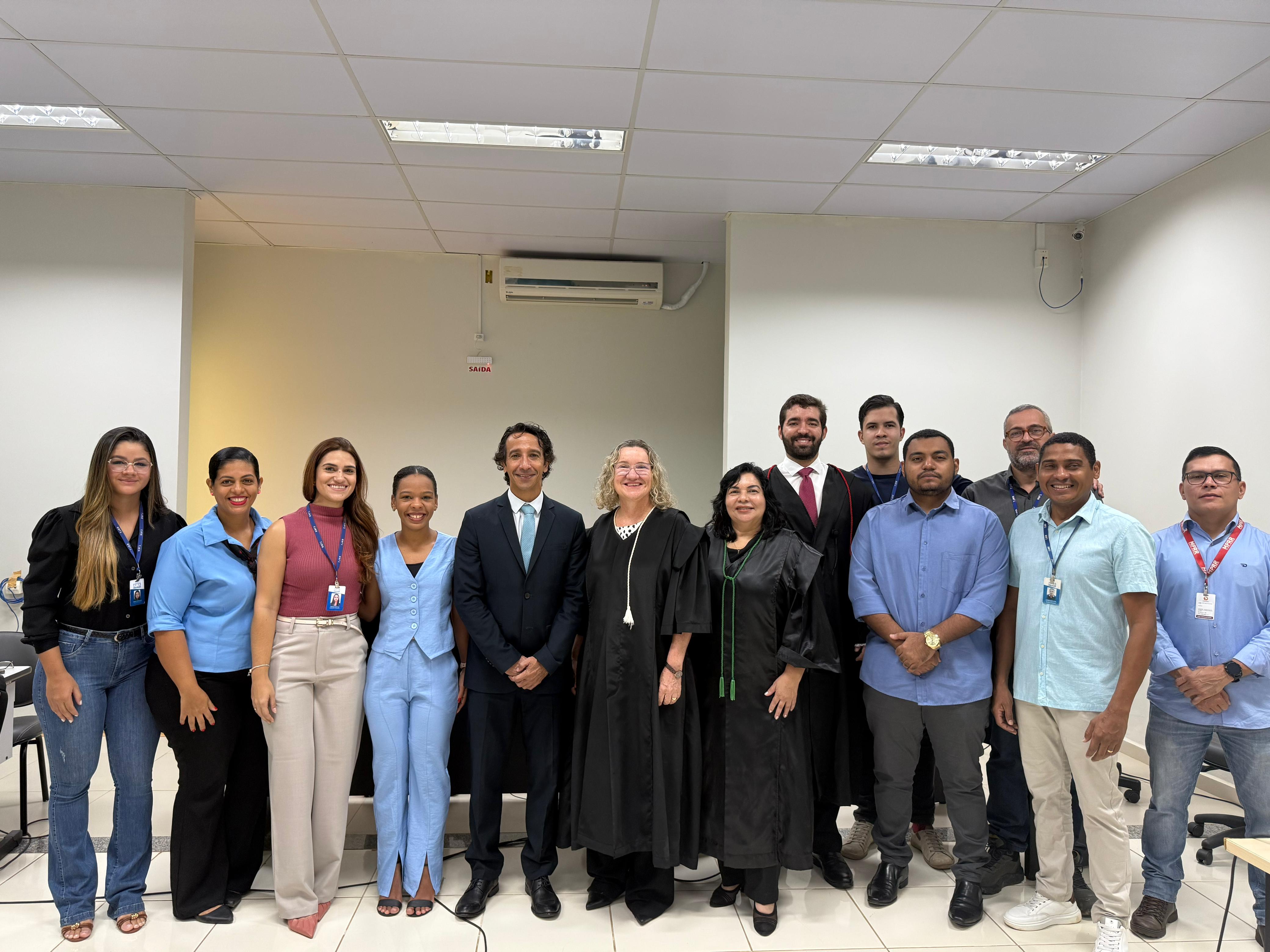 Grupo de 13 pessoas posa sorridente para foto em uma sala com iluminação clara e teto de gesso. Ao centro, três magistrados usam toga preta, cercados por servidores e colaboradores com crachás e roupas formais. O ambiente é de um local institucional, com ar-condicionado visível na parede ao fundo. A atmosfera é de celebração ou encerramento de atividade oficial. Grupo de 13 pessoas posa sorridente para foto em uma sala com iluminação clara e teto de gesso. Ao centro, três magistrados usam toga preta, cercados por servidores e colaboradores com crachás e roupas formais. O ambiente é de um local institucional, com ar-condicionado visível na parede ao fundo. A atmosfera é de celebração ou encerramento de atividade oficial.