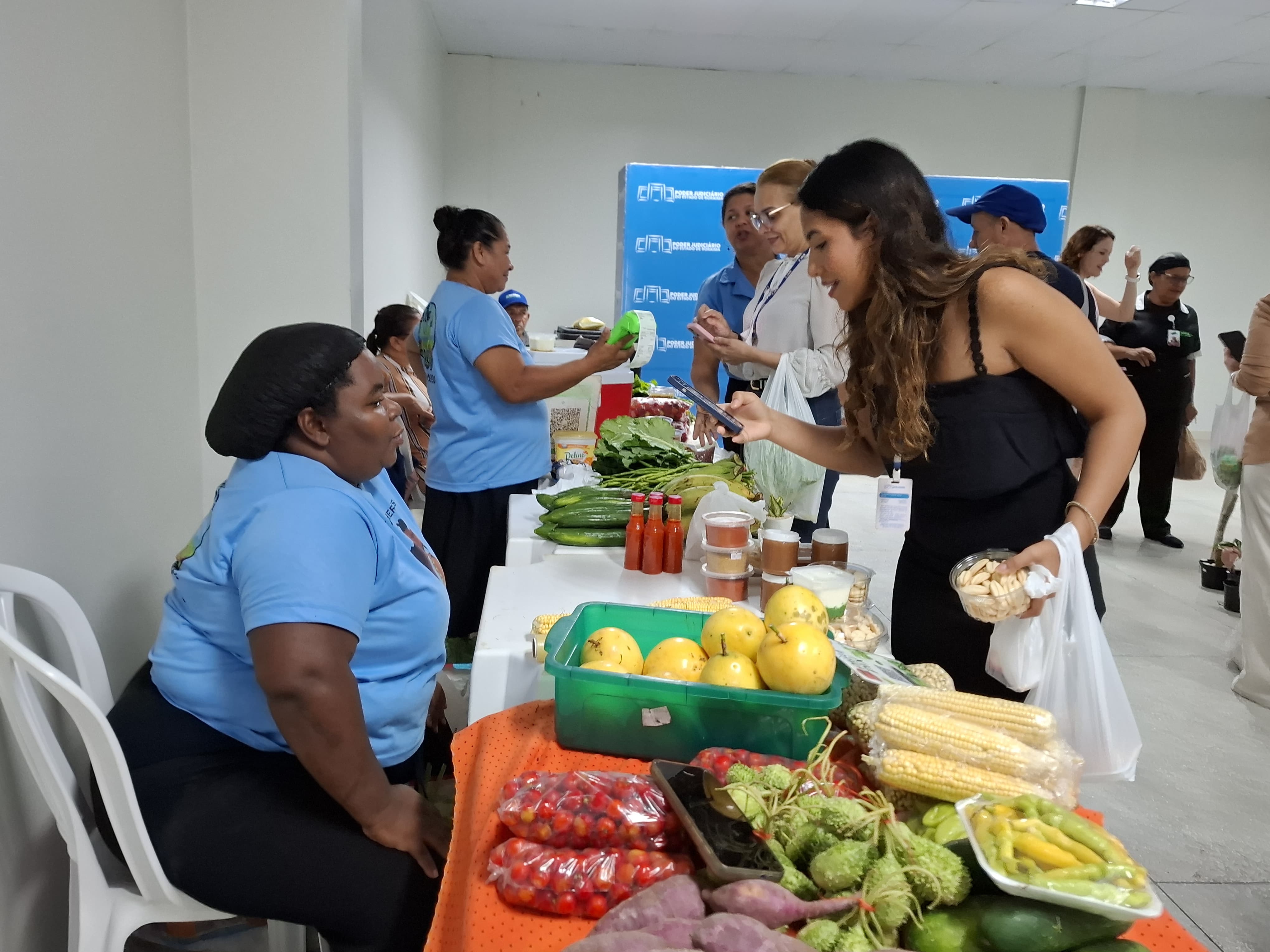 Foto colorida mostra um corredor movimentado da feira com várias pessoas comprando e conversando. Em primeiro plano, uma vendedora sentada atende uma visitante que observa os produtos na mesa. Há frutas como maracujá, milho, batata-doce, maxixe e embalagens com doces ou compotas. Foto colorida mostra um corredor movimentado da feira com várias pessoas comprando e conversando. Em primeiro plano, uma vendedora sentada atende uma visitante que observa os produtos na mesa. Há frutas como maracujá, milho, batata-doce, maxixe e embalagens com doces ou compotas.