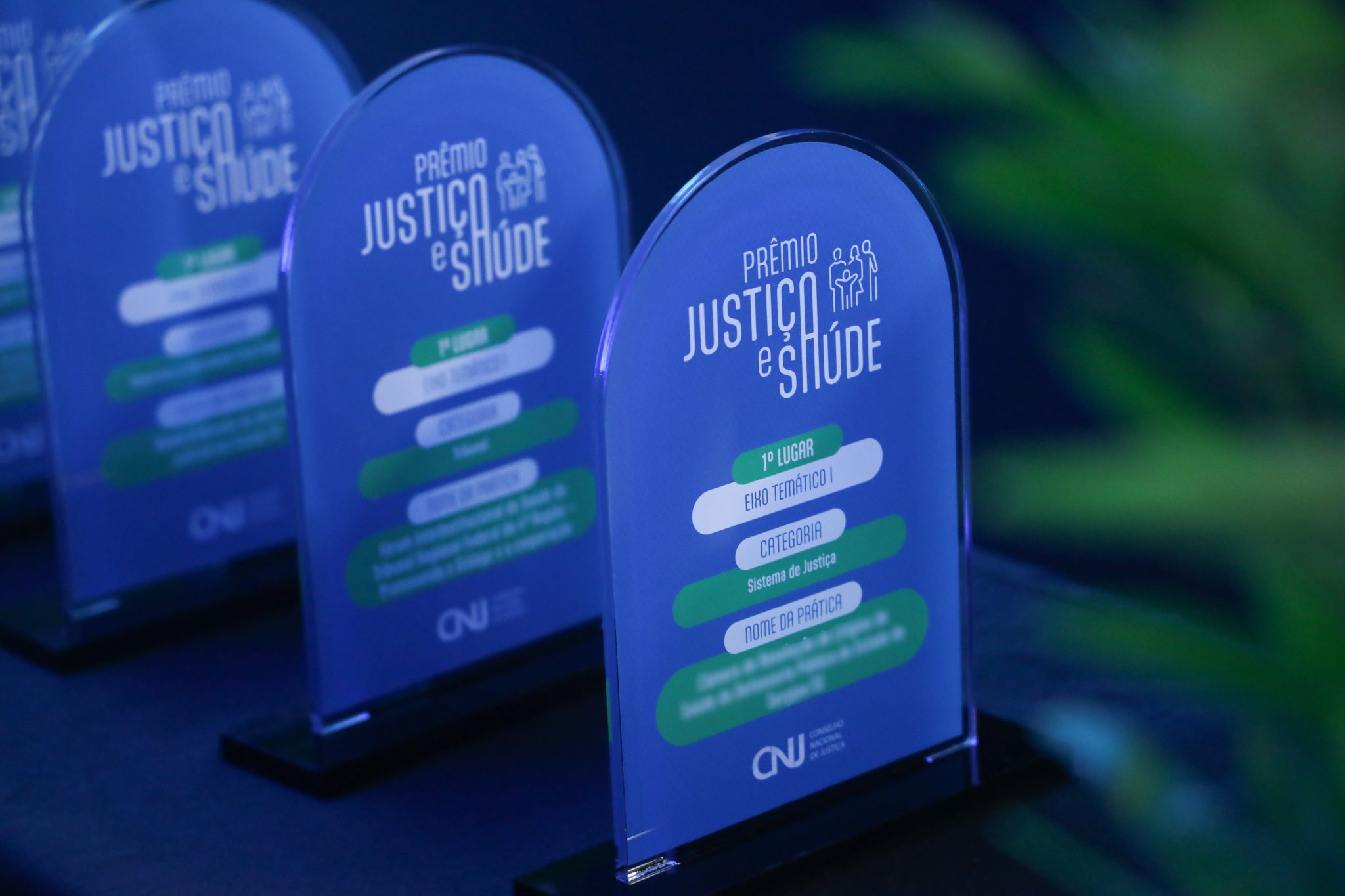 Foto colorida mostra troféus do Prêmio Justiça e Saúde, com estrutura azul em acrílico e base preta. Em destaque, a placa do 1º lugar com informações sobre eixo temático, categoria e nome da prática. O logotipo do Conselho Nacional de Justiça (CNJ) aparece na parte inferior da premiação. Foto colorida mostra troféus do Prêmio Justiça e Saúde, com estrutura azul em acrílico e base preta. Em destaque, a placa do 1º lugar com informações sobre eixo temático, categoria e nome da prática. O logotipo do Conselho Nacional de Justiça (CNJ) aparece na parte inferior da premiação.
