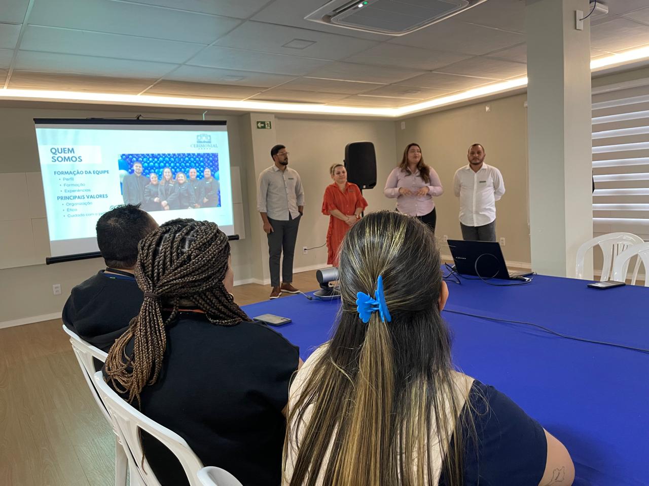 Imagem colorida em ambiente interno com a turma sentada. À esquerda, uma tela grande exibe uma apresentação com texto que inclui "QUEM SOMOS" e "FORMAÇÃO DA EQUIPE" Uma mulher com uma blusa rosa claro está falando. Um grupo de participantes está sentado em uma longa mesa em primeiro plano, de frente para os apresentadores e a tela. A mesa está coberta com um pano azul.