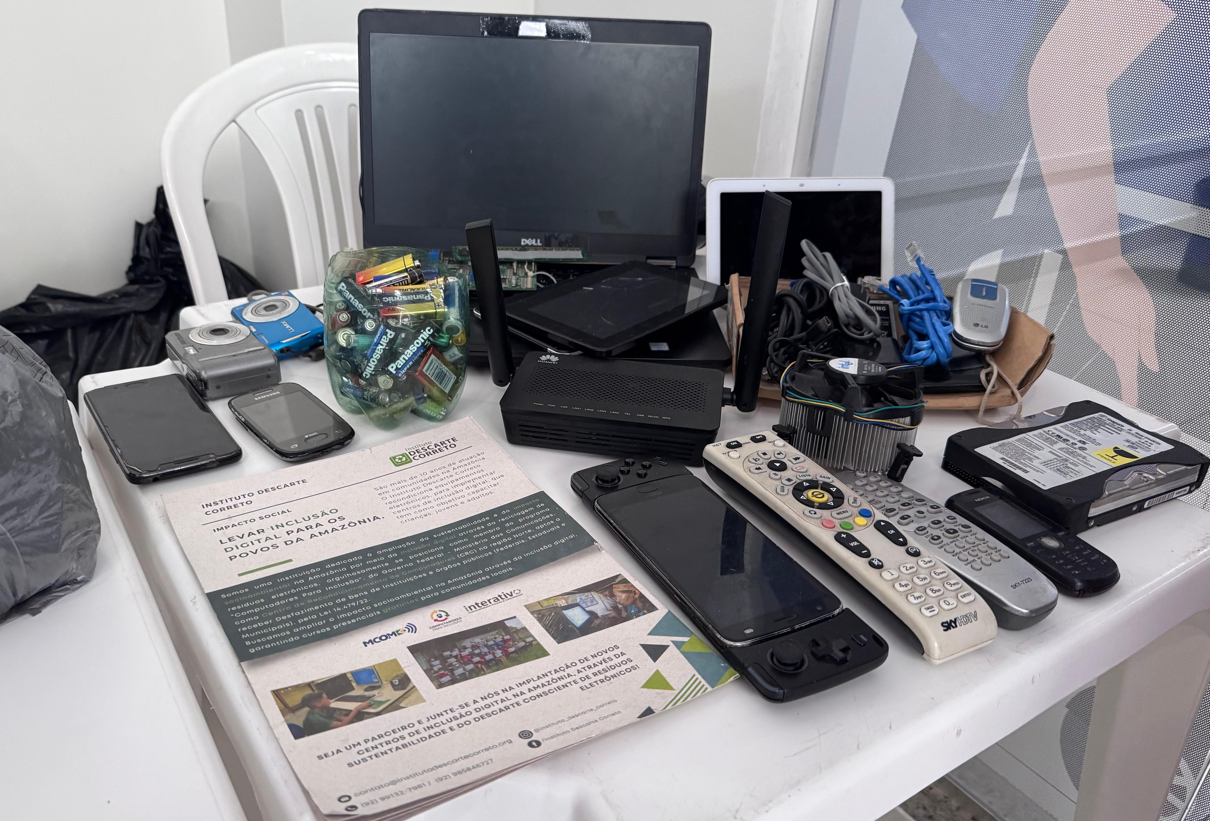  Imagem colorida mostra uma mesa branca coberta com diversos aparelhos eletrônicos descartados, como notebooks, tablets, pilhas, celulares, controles remotos e cabos.