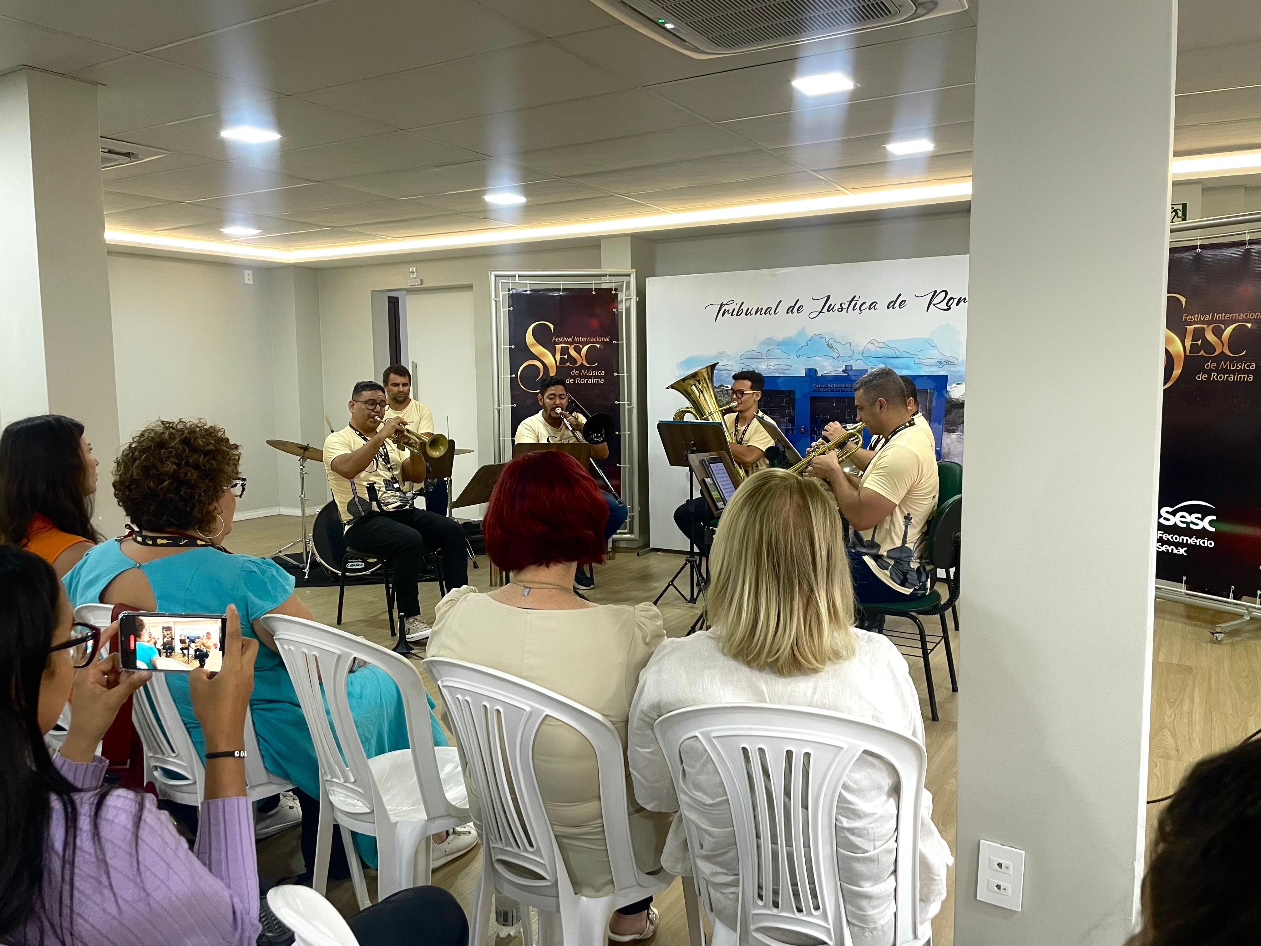 Imagem colorida mostra a plateia sentada em cadeiras brancas, de costas para a câmera, assistindo aos músicos no palco. Quatro músicos, vestidos com camisetas bege, estão tocando instrumentos de sopro. Um quinto músico, visível à esquerda, está tocando bateria. Ao fundo, o mesmo banner do "Tribunal de Justiça de Roraima" e o banner do "Festival Internacional SESC de Música de Roraima" são visíveis.