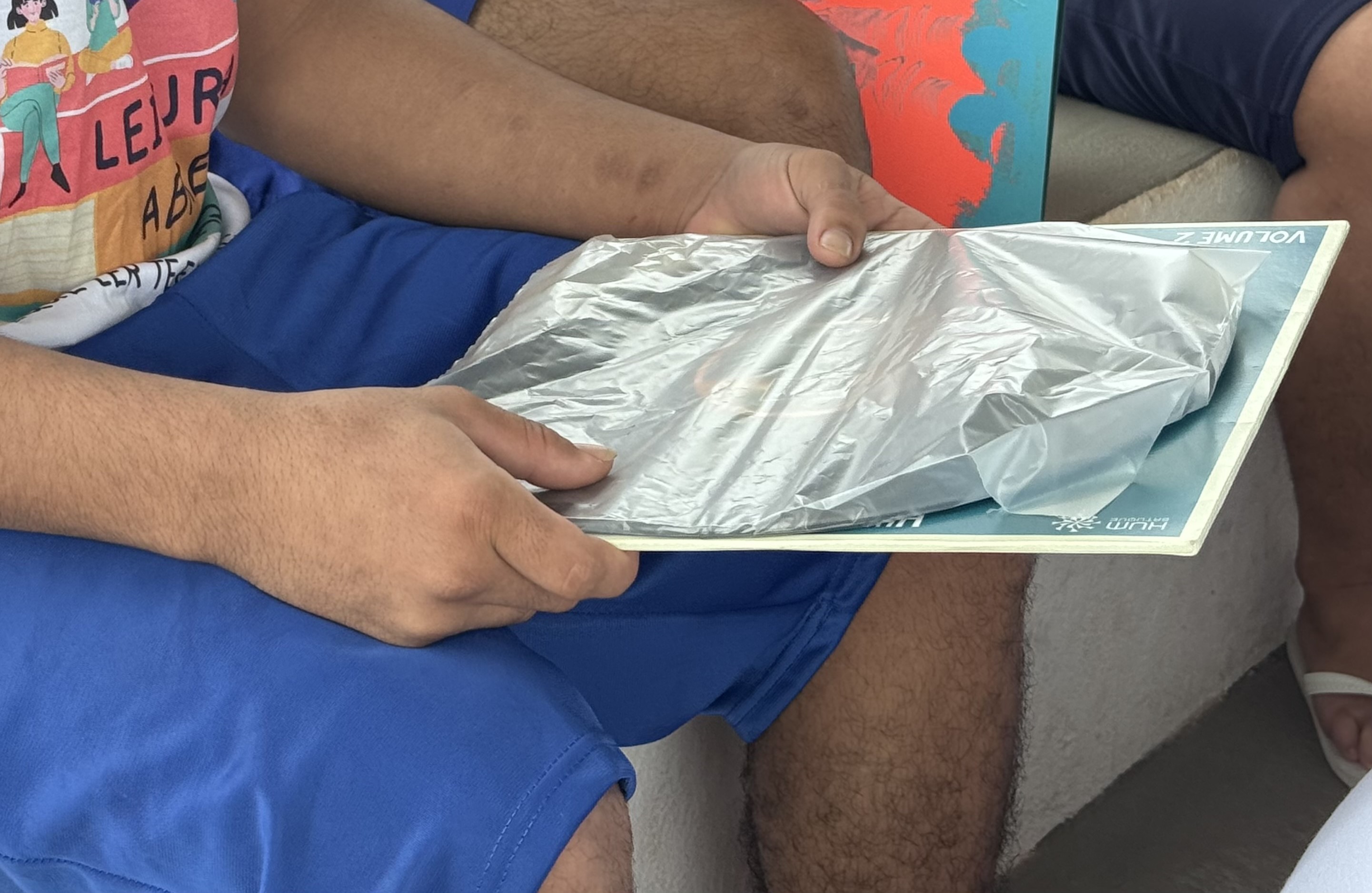 Imagem colorida mostra uma pessoa segurando um disco de vinil coberto por plástico. A pessoa veste um calção azul e parte de uma camiseta branca. Outra pessoa e um objeto colorido são visíveis ao fundo.