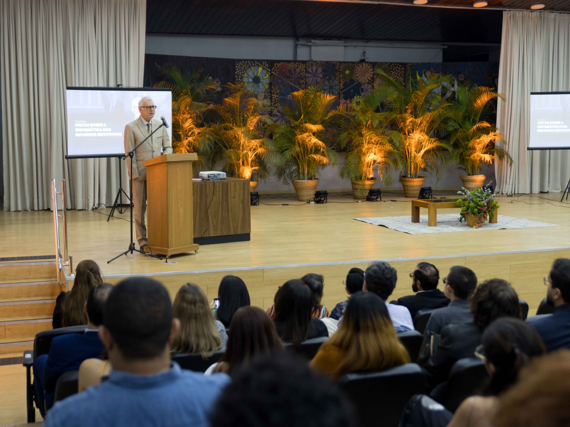 magem colorida do ministro no púlpito de madeira em pé, vestido de terno claro e óculos, falando ao microfone. No fundo, há uma parede com um mural colorido e cortinas bege. Ao lado do palco, há vasos de plantas tropicais. Na plateia, as pessoas estão sentadas, assistindo a palestra. magem colorida do ministro no púlpito de madeira em pé, vestido de terno claro e óculos, falando ao microfone. No fundo, há uma parede com um mural colorido e cortinas bege. Ao lado do palco, há vasos de plantas tropicais. Na plateia, as pessoas estão sentadas, assistindo a palestra.