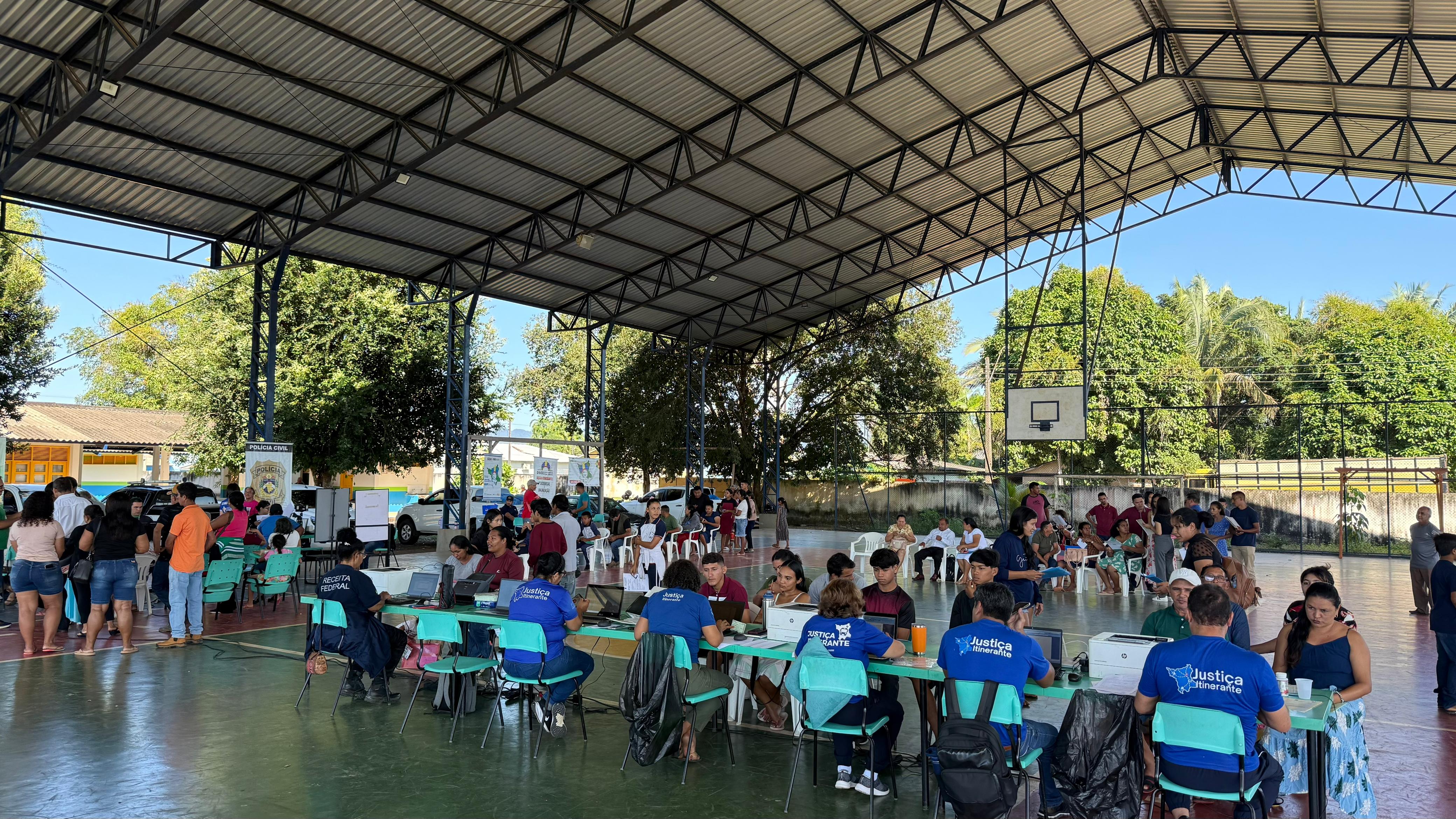 Foto colorida mostra um evento de atendimento público social sendo realizado em uma quadra poliesportiva coberta. A quadra tem o chão de cimento pintado de verde. Diversas mesas estão dispostas em filas, com atendentes usando camisetas azuis com a logo "Justiça Itinerante" visível. O público, composto por adultos e crianças, aguarda em cadeiras ou em pé nas filas. Ao fundo, é possível ver árvores e o muro de uma escola.