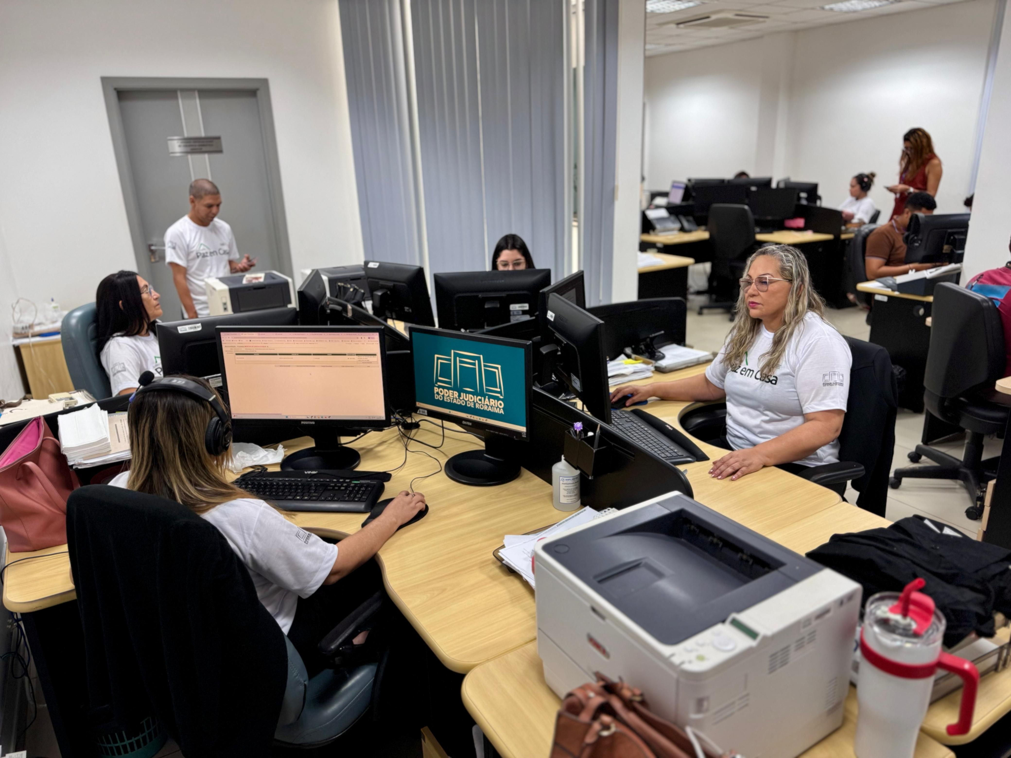 imagem mostra um ambiente de trabalho dando enfoque em cinco pessoas, a maioria mulheres, em suas estações de trabalho.  Todos estão vestindo blusas brancas escrito "Paz em casa"
