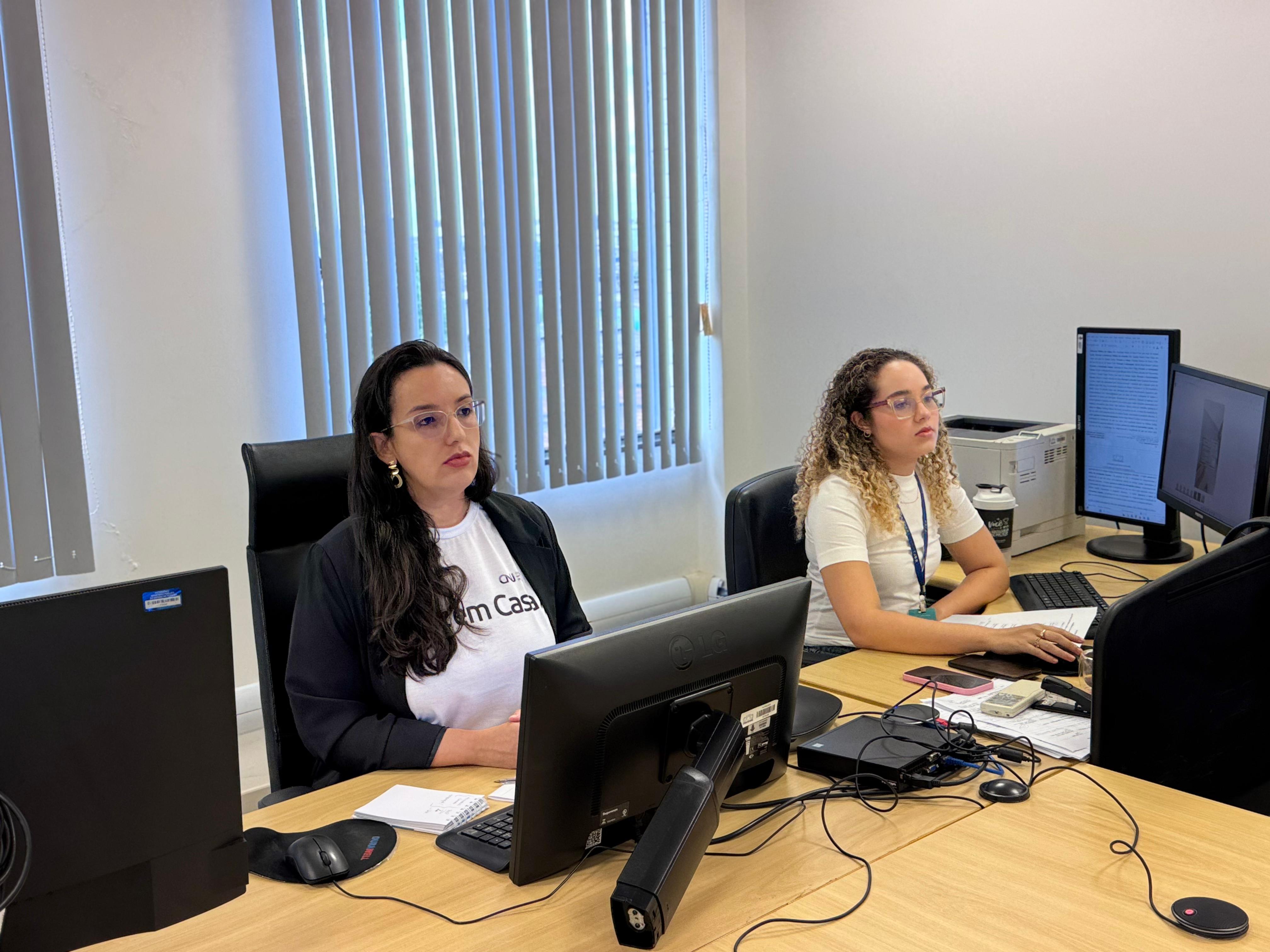 imagem mostra um ambiente de trabalho dando enfoque em duas pessoas, duas mulheres que estão em suas estação de trabalho. Uma está vestindo blusas brancas escrito "Paz em casa".