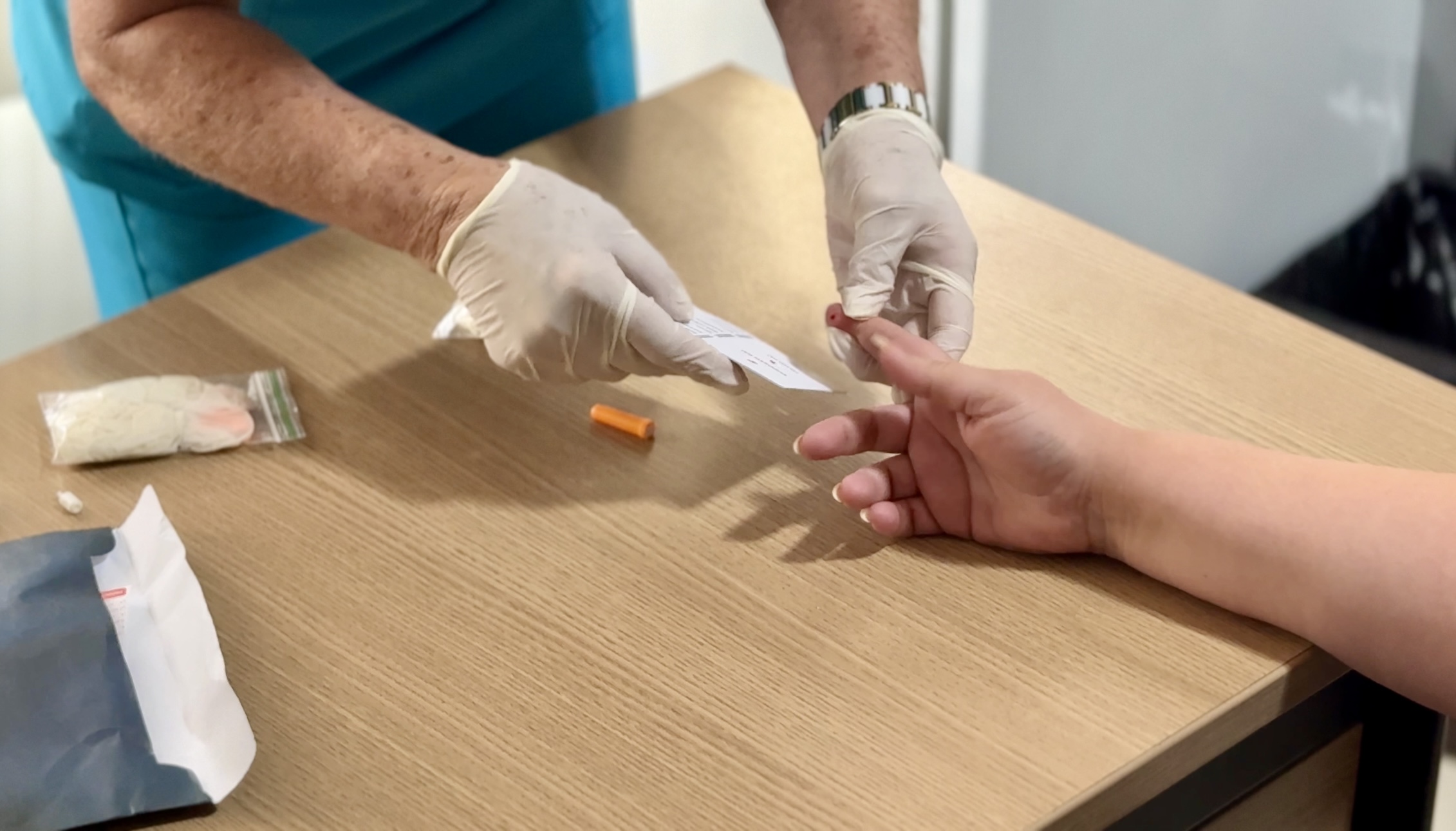 Imagem colorida mostra as mãos de uma pessoa de avental azul e luvas brancas, perfurando o dedo de outra pessoa para coleta de sangue. Um pequeno papel branco com um código de barras está sendo segurado próximo ao dedo. Um kit para teste, incluindo uma bandagem e um pequeno tubo laranja, está sobre a mesa de madeira clara.