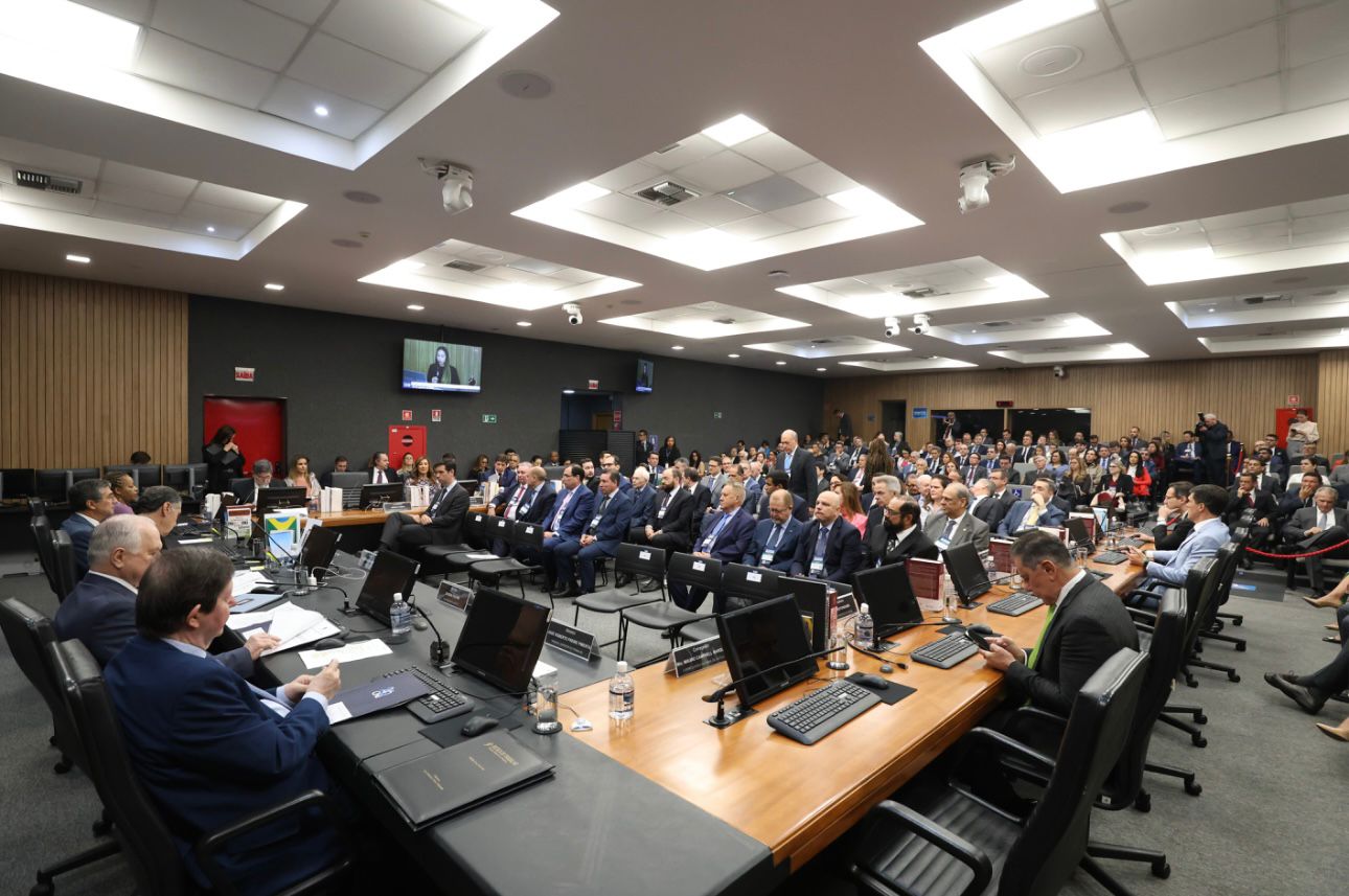  Imagem colorida mostra uma sala de conferências grande, com muitas pessoas sentadas em fileiras de cadeiras e outras em uma mesa comprida. A maioria das pessoas está vestindo ternos. Há computadores, notebooks e garrafas de água sobre a mesa principal. No centro da sala, um telão mostra uma mulher em um vídeo.