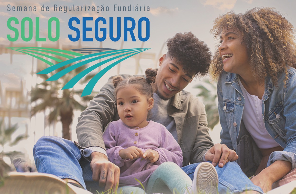 uma foto de uma família sorrindo, sentada na grama. Um homem de cabelo crespo e barba rala, uma mulher de cabelo cacheado e uma criança pequena de cabelo preso em dois coques. Eles olham para o lado direito da foto. A mulher e o homem seguram as mãos da criança. No topo da foto há um texto com a frase Semana de Regularização Fundiária e logo abaixo o slogan SOLO SEGURO, com um desenho de linhas curvas em tons de azul e verde que se assemelham a um campo de terra.