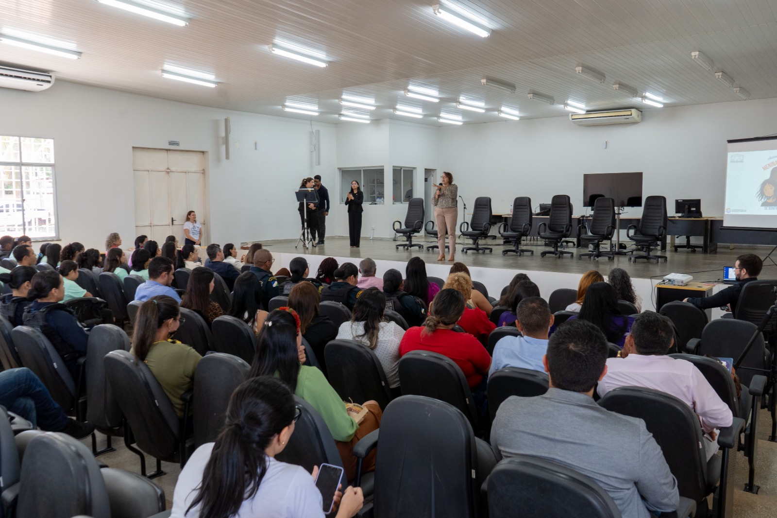 fotografia tirada de frente, mostra um grande auditório com um público em cadeiras pretas assistindo a uma palestra. No palco, mais elevado, há um painel de cadeiras vazias à direita e, ao centro, três pessoas em pé, sendo uma delas uma mulher falando a um microfone. fotografia tirada de frente, mostra um grande auditório com um público em cadeiras pretas assistindo a uma palestra. No palco, mais elevado, há um painel de cadeiras vazias à direita e, ao centro, três pessoas em pé, sendo uma delas uma mulher falando a um microfone.