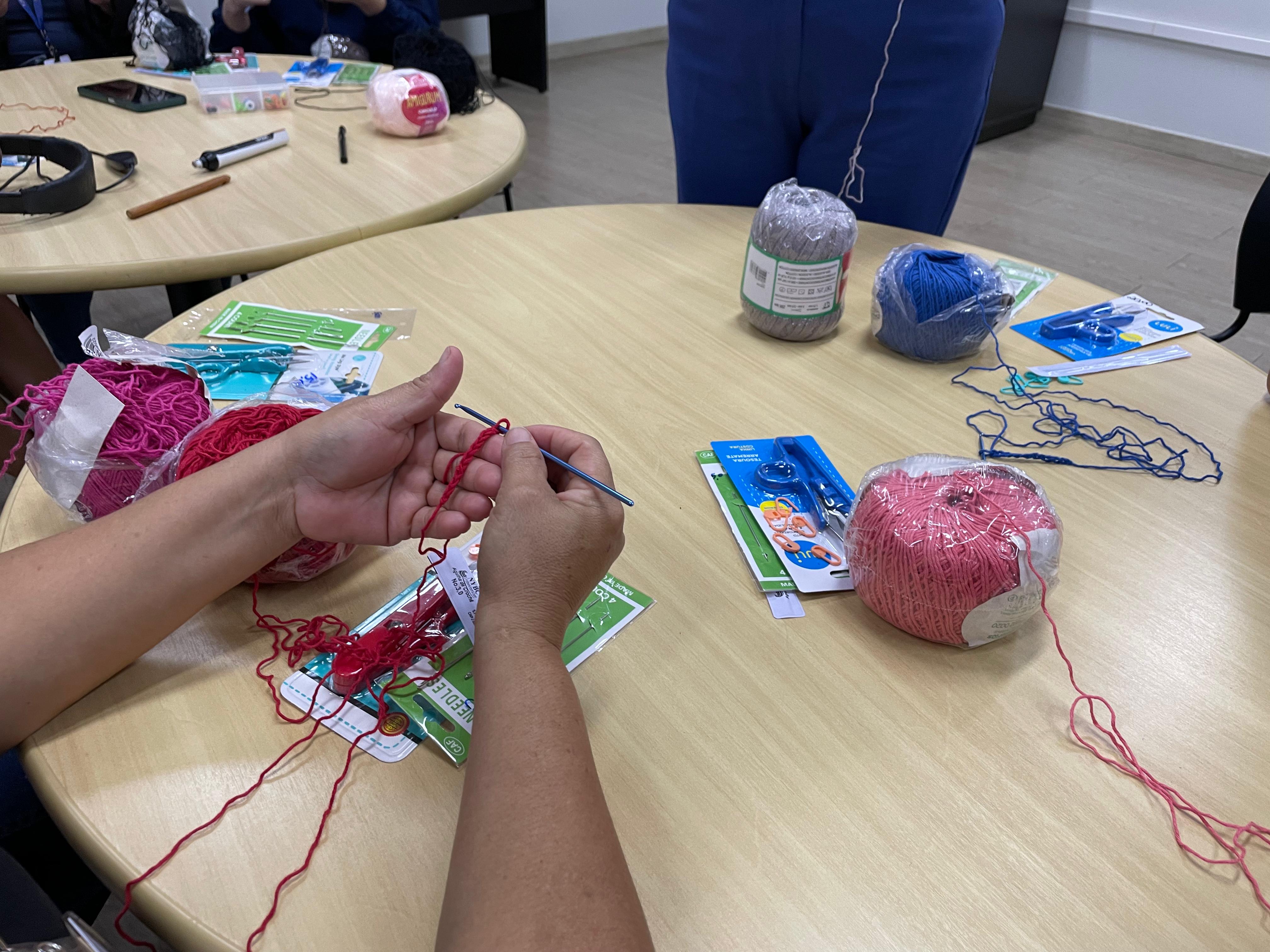 Imagem colorida mostra um close de mãos fazendo crochê com uma linha vermelha. Sobre a mesa, há novelos de lã de diferentes cores (vermelho, rosa, azul, cinza), embalagens de agulhas e outras ferramentas para a atividade. Ao fundo, pode-se ver parte de uma mesa redonda e pernas de pessoas sentadas.