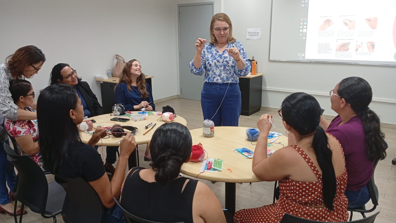 Imagem colorida mostra a  Presidente da Comissão, Desembargadora Elaine Bianchi usando uma blusa florida e calça azul, que está em pé, no centro de uma sala, ensinando um grupo de mulheres a fazer crochê. As alunas estão sentadas ao redor de duas mesas redondas, com linhas e agulhas. Há uma lousa digital ao fundo, exibindo instruções de crochê com imagens.