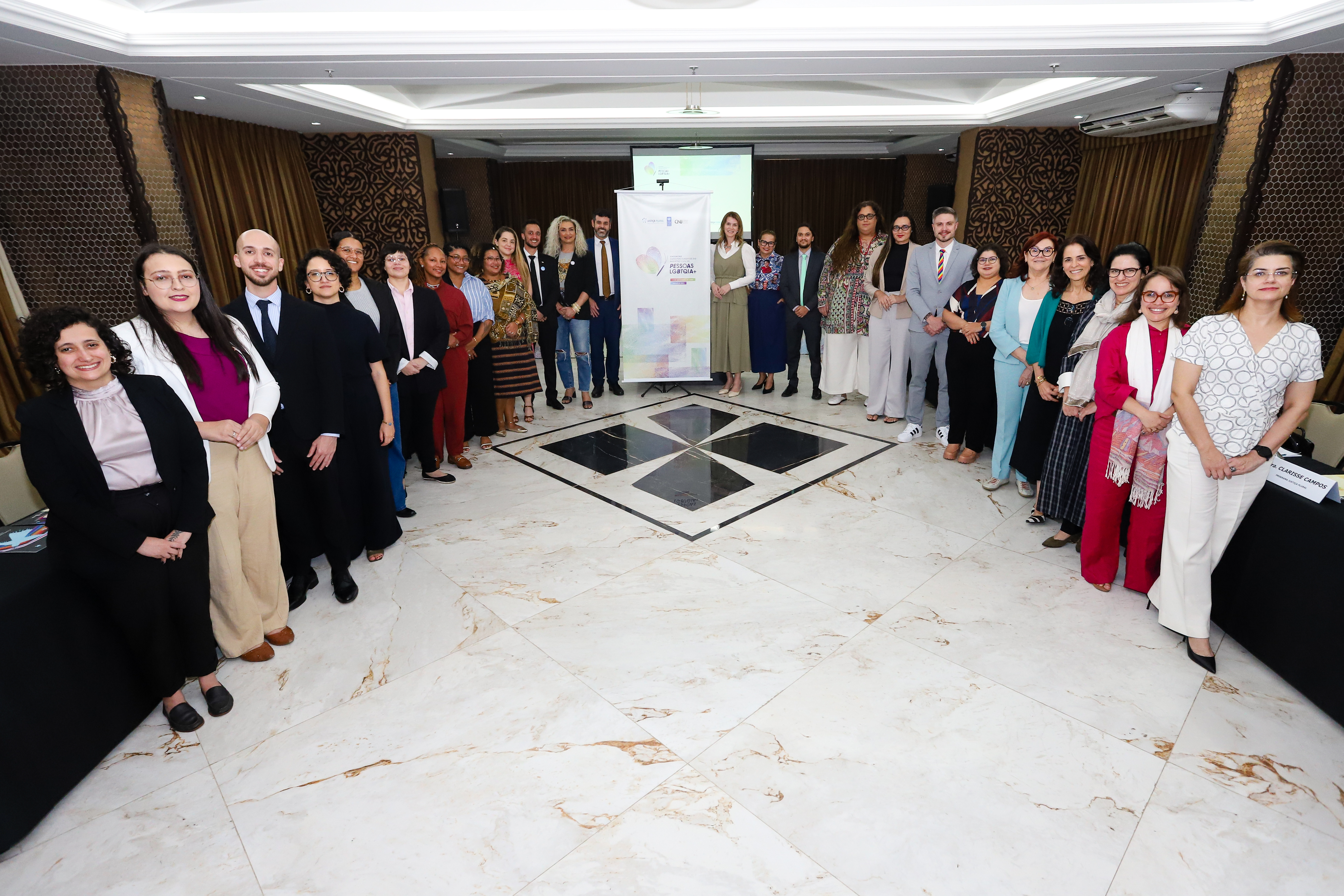 Foto mostra um grupo diverso de pessoas posando para a foto em uma sala ampla e bem iluminada, com piso de mármore claro e cortinas escuras ao fundo. Os participantes estão dispostos em duas fileiras laterais, formando um corredor central que leva a um banner institucional posicionado ao fundo. Durante sua participação, a magistrada ressaltou o mutirão “Trans‑Formando Identidades”, iniciativa da Justiça Itinerante em parceria com a Defensoria Pública de Roraima (DPE-RR), que busca facilitar o acesso ao registro civil e garantir direitos fundamentais dessa população.