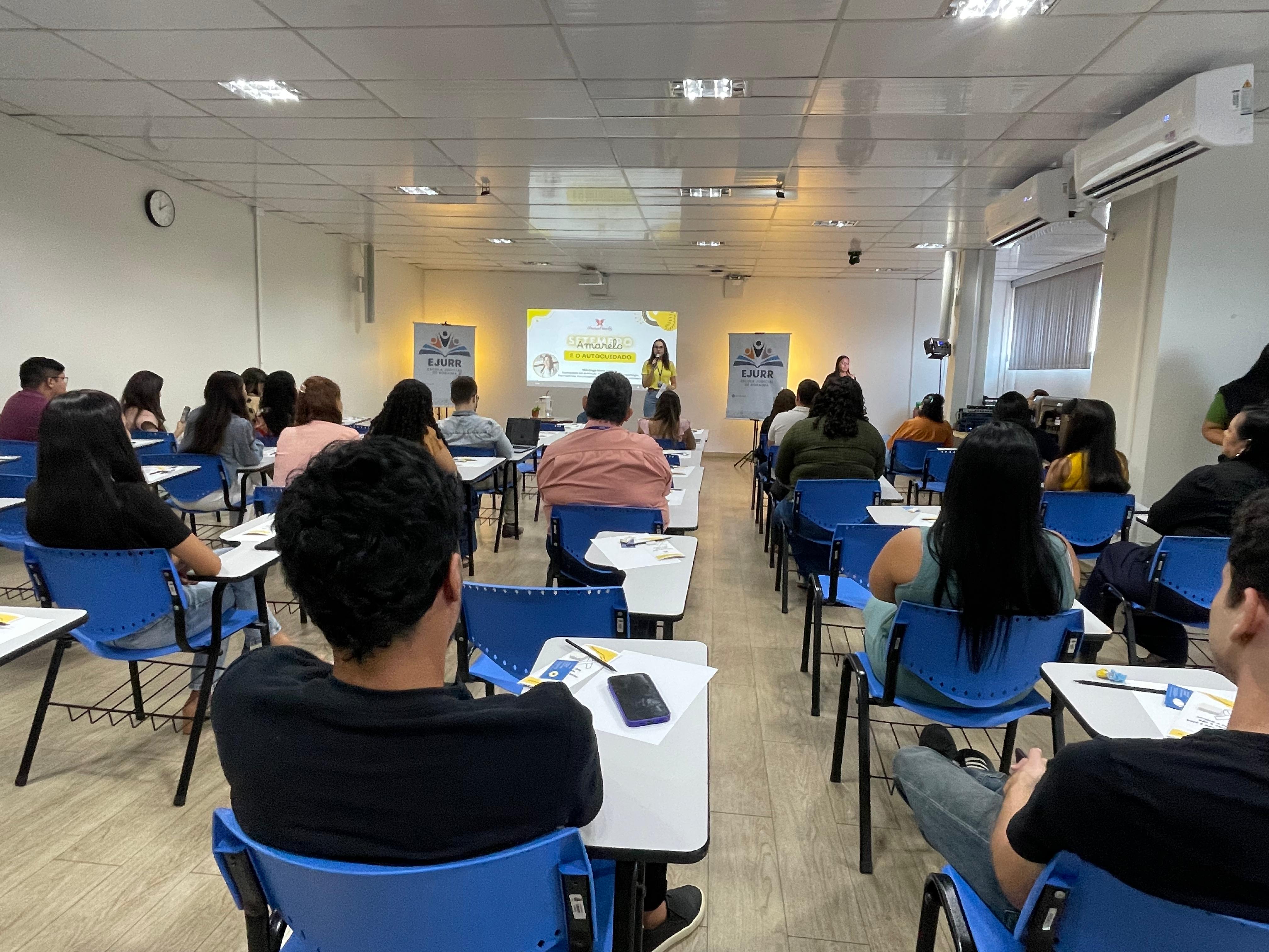 Foto colorida mostra uma sala, com participantes sentados em cadeiras escolares azuis, voltados para o fundo da sala, onde ocorre uma palestra. No centro do ambiente, uma mulher vestindo amarelo fala ao microfone, enquanto outra pessoa a acompanha ao lado. O telão exibe a temática “Setembro Amarelo e o Autocuidado”