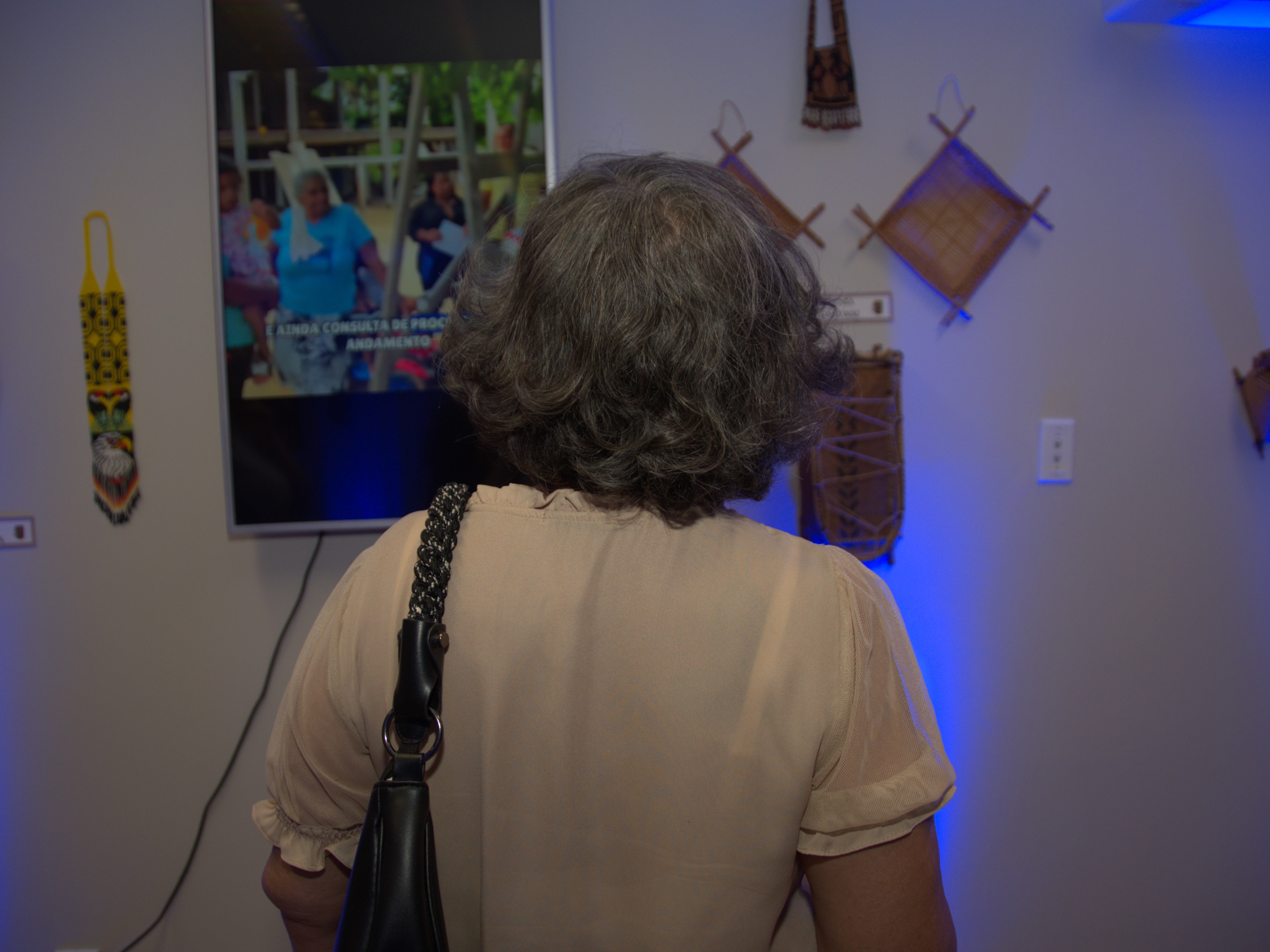Imagem mostra uma mulher, vista de costas, observando atentamente uma tela de televisão em exposição. A TV exibe uma cena com pessoas, enquanto a parede ao redor está decorada com artesanatos indígenas, incluindo cestos e peças trançadas. 