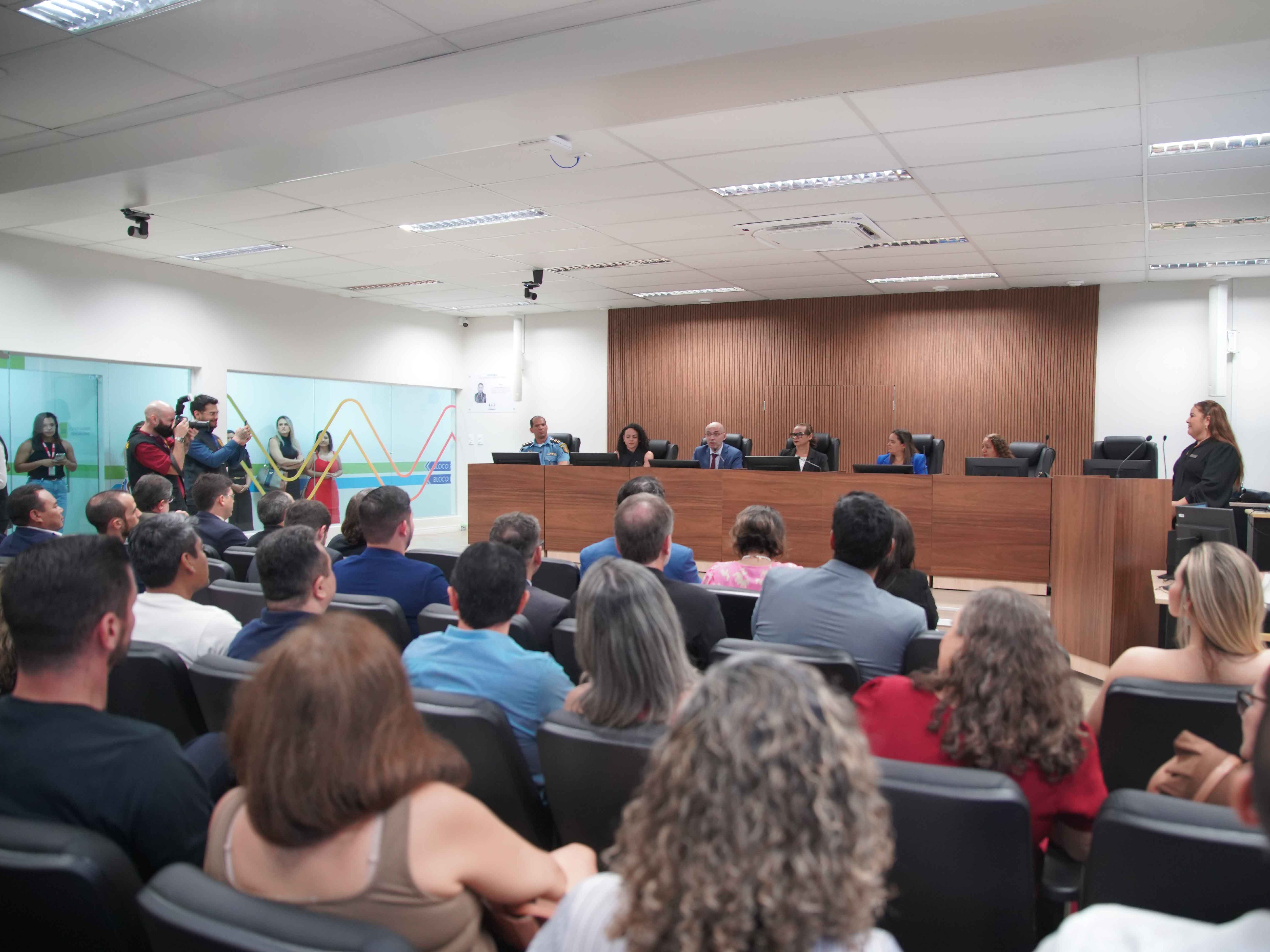 Imagem mostra um auditório cheio, com público sentado de frente para uma mesa de autoridades composta por seis pessoas, entre homens e mulheres, em um ambiente moderno com painéis de madeira ao fundo. À esquerda, fotógrafos e convidados registram o momento; à direita, uma mulher em pé fala ao microfone. A sala tem iluminação clara, ar-condicionado e cadeiras pretas.
